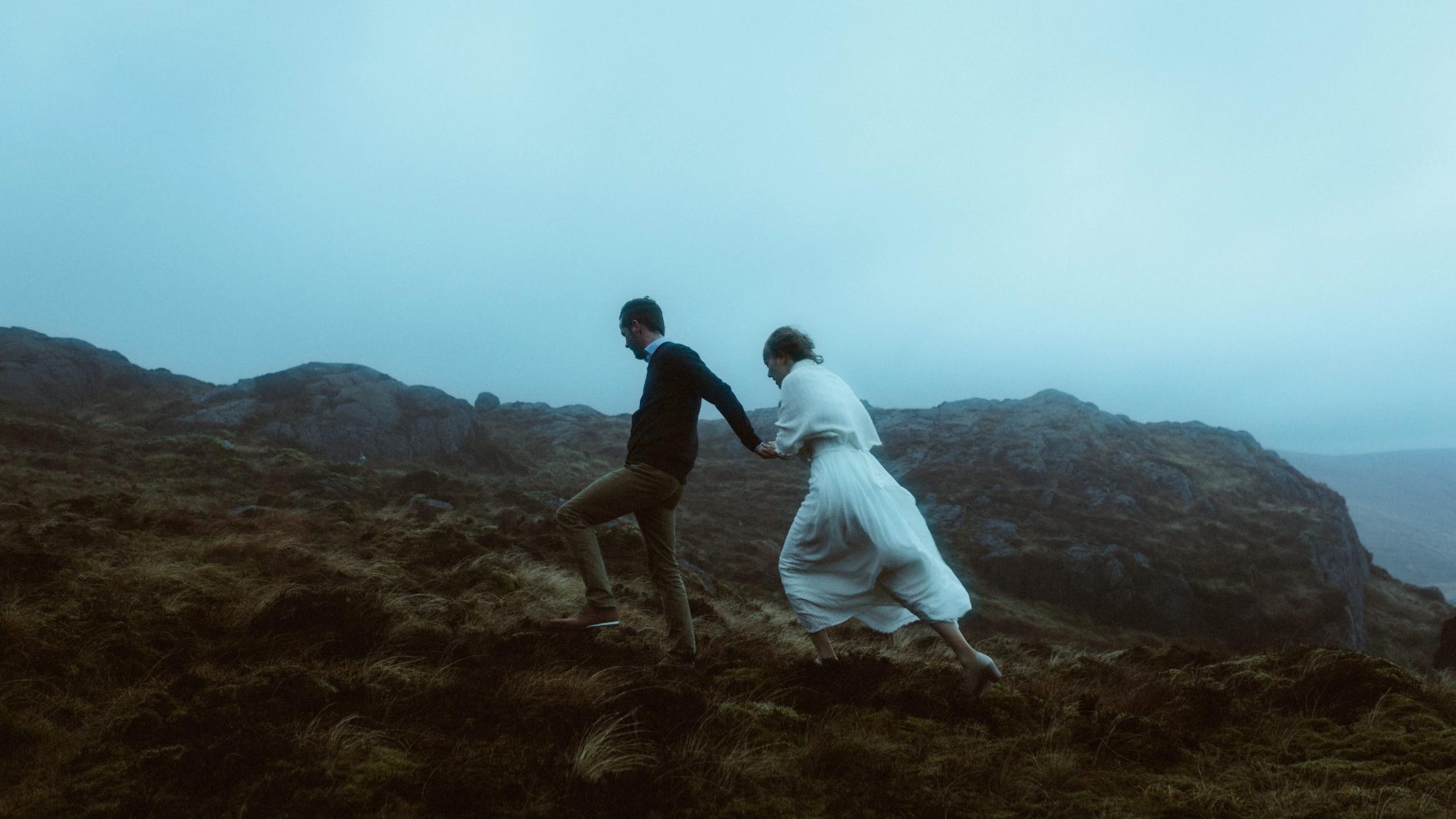 Jule und Rory laufen im Nebel bergauf, ihr weißes Kleid wird vom Wind bewegt.