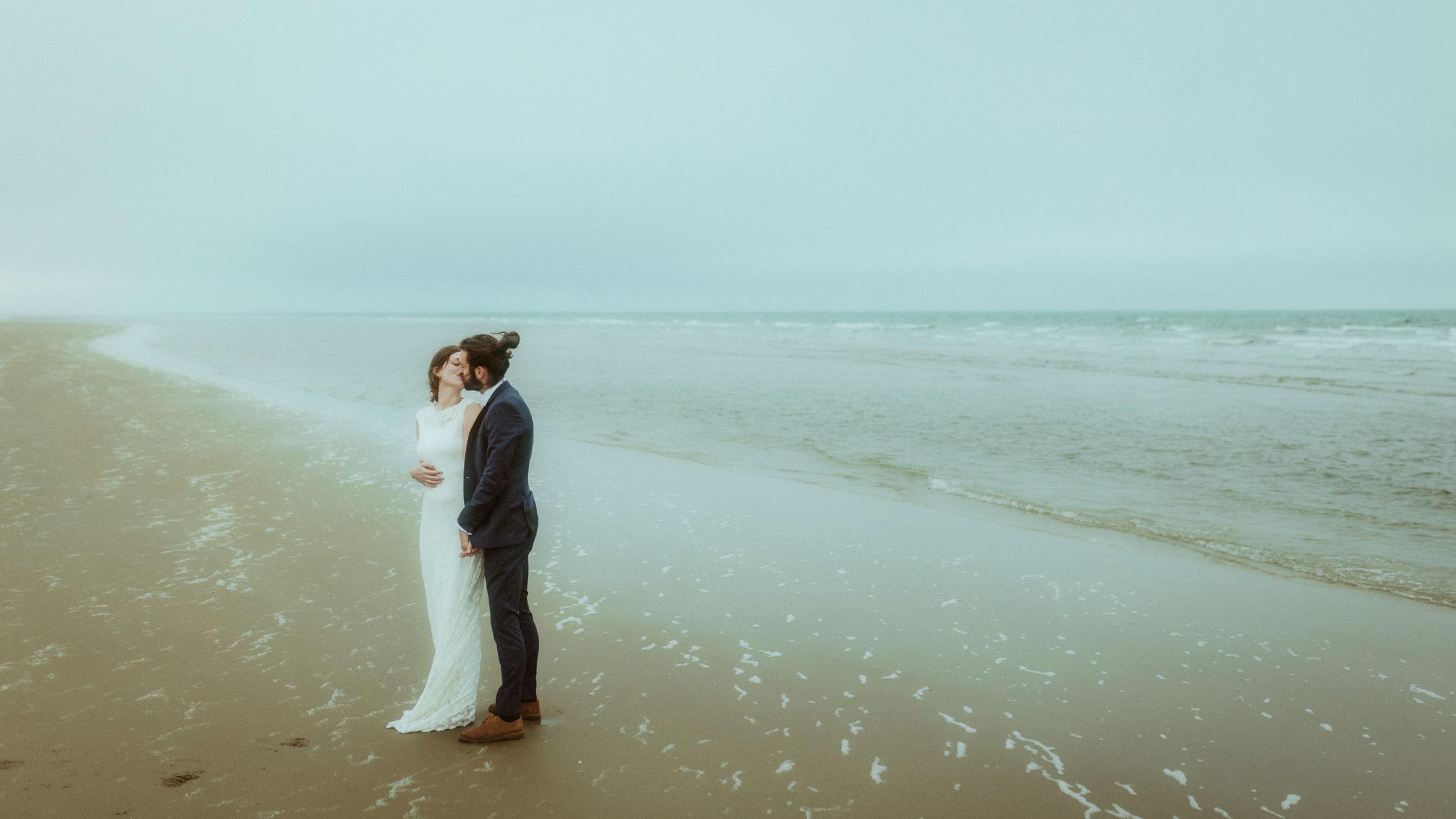 Nina und Torben umarmen sich am leeren Strand, hinter ihnen erstreckt sich das Meer.
