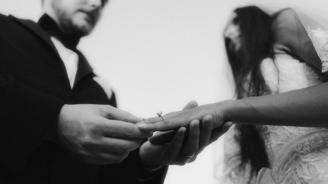 In a black-and-white close-up, the groom holds the bride's hand and touches her ring.