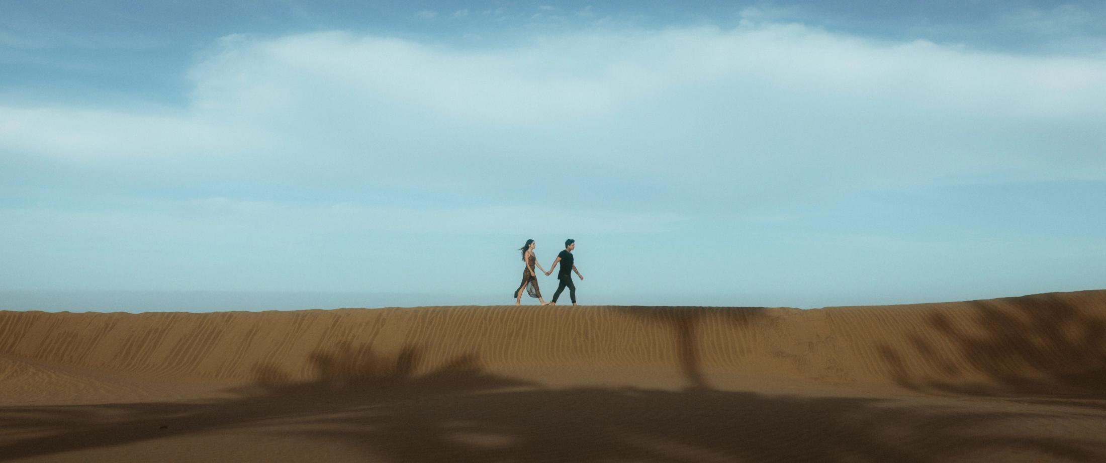 Weitwinkelaufnahme, Angela und Ivan gehen Hand in Hand über einen Dünenkamm, klein vor einem riesigen Himmel.