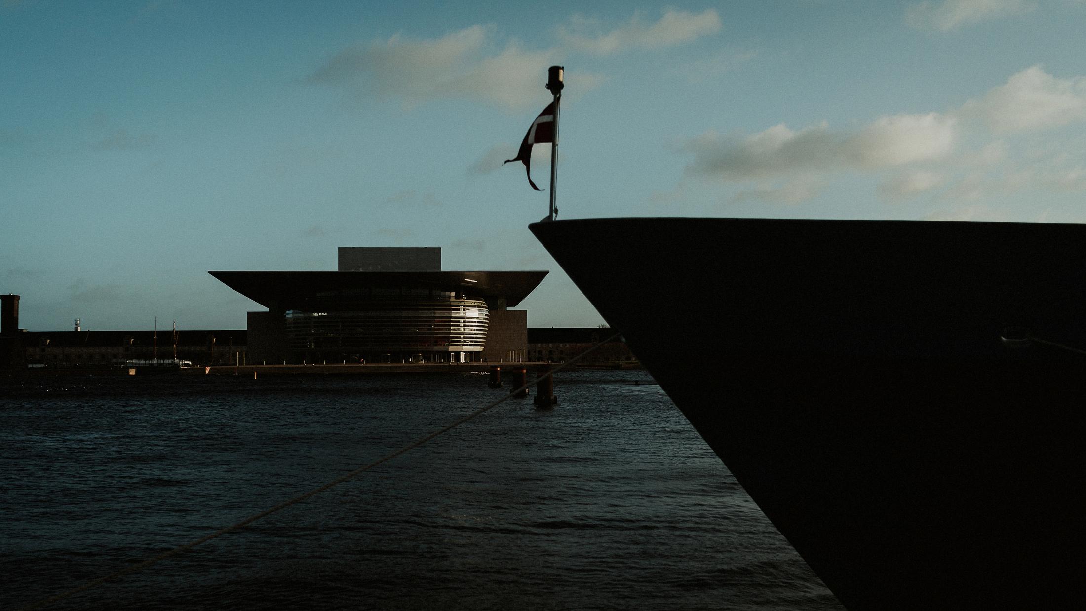 A dark ship’s bow frames the Copenhagen Opera House across the water.