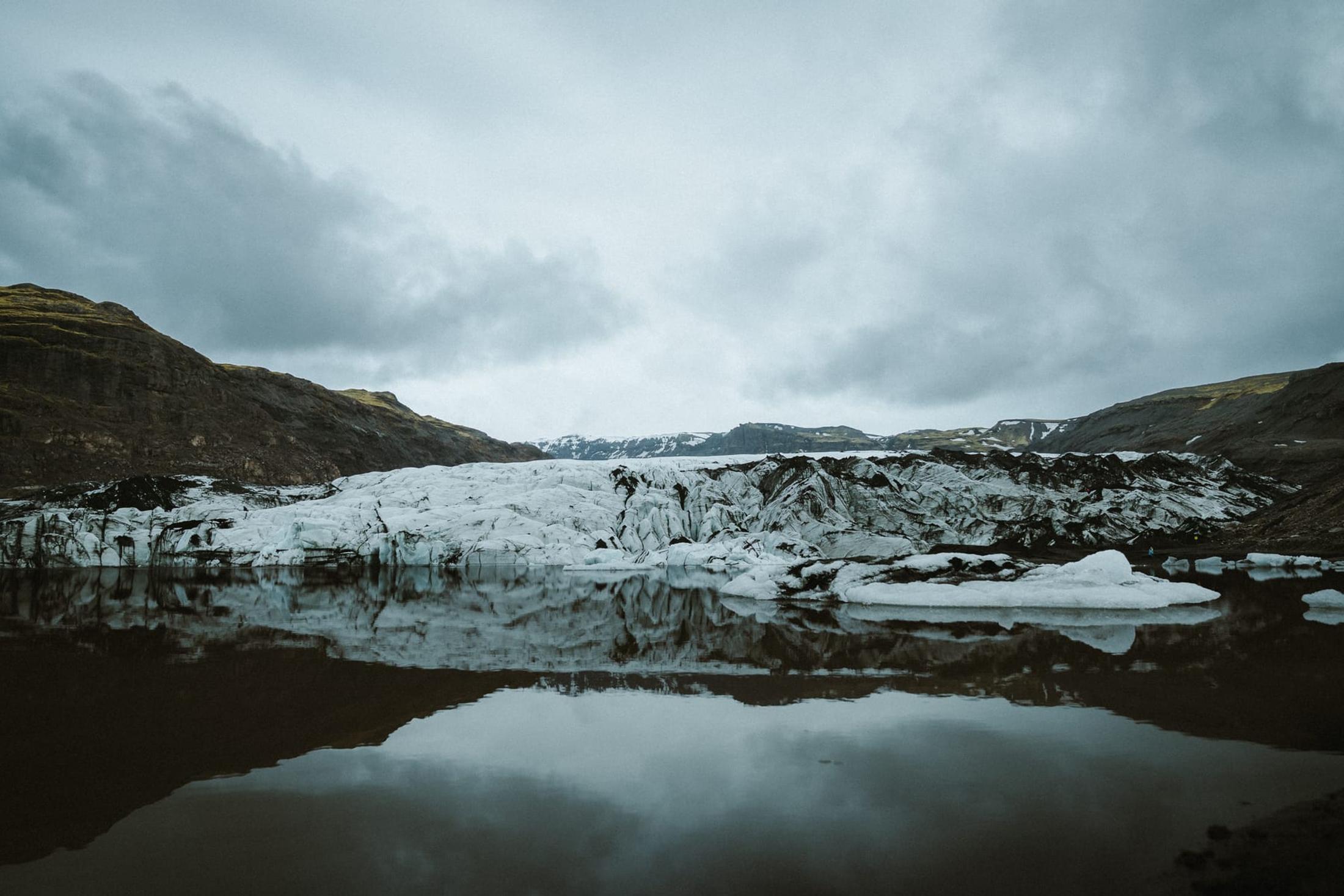 A glacier tongue reflected in still water, between dark slopes beneath an overcast sky.