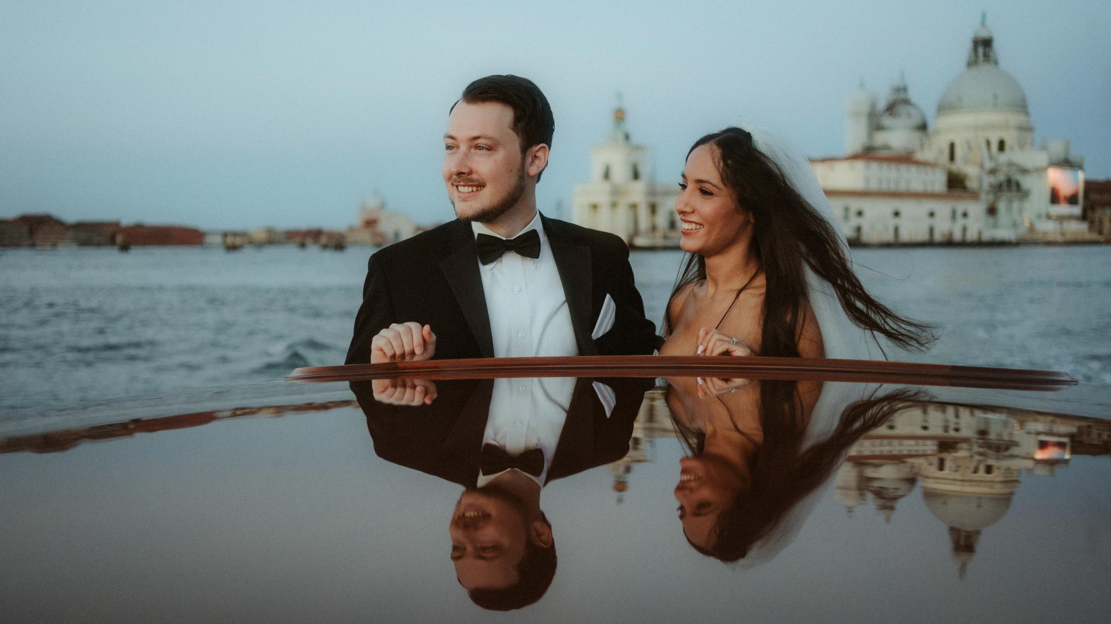 The couple laughs on a boat, mirrored in the polished wooden surface in front of them.
