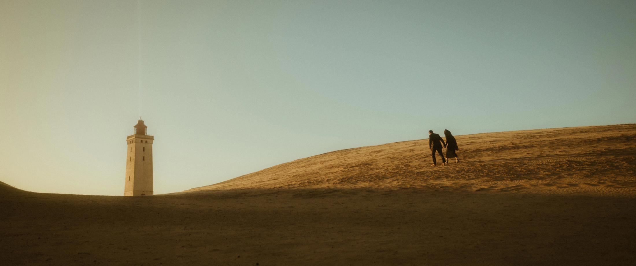 A couple walks as small silhouettes through the dunes toward a lighthouse.
