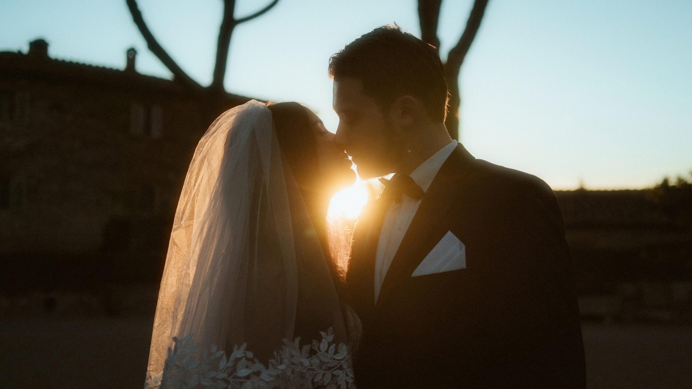 The couple kisses with the low evening sun shining between their faces.