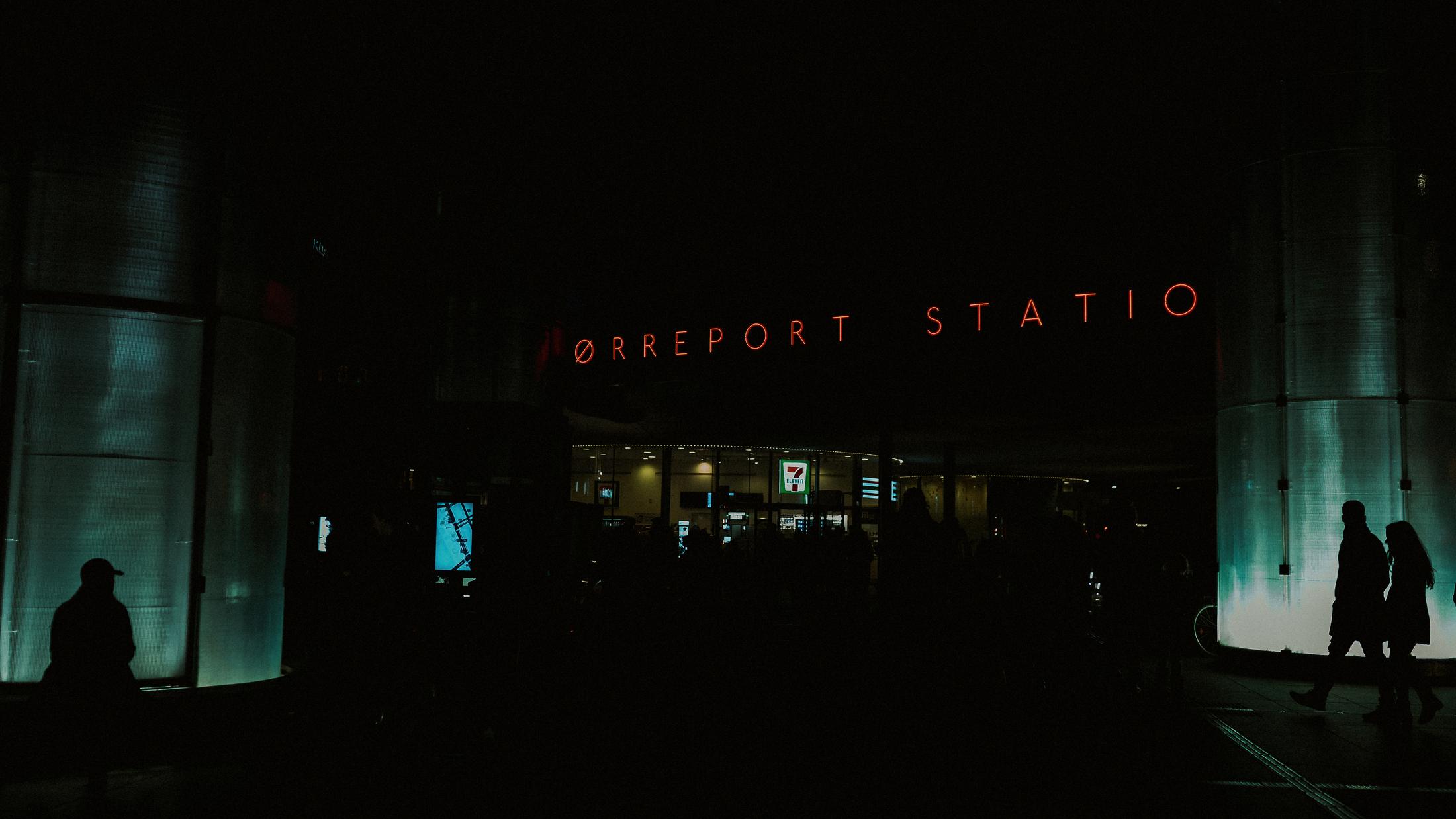 Pedestrians appear in silhouette beneath the red Nørreport Station sign at night.