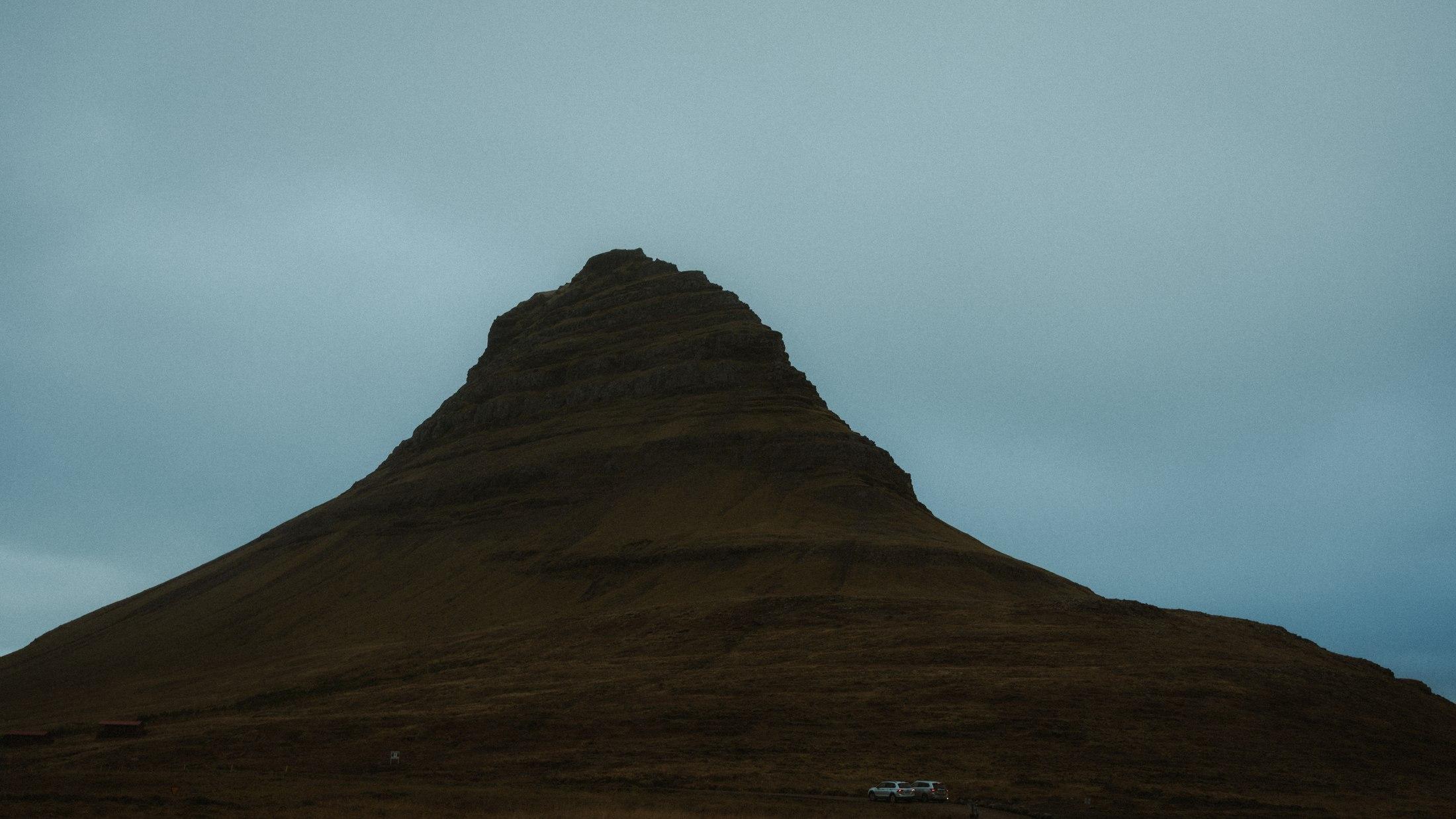 The layered slopes of Kirkjufell beneath a grey-blue sky, with cars at its base showing the scale.