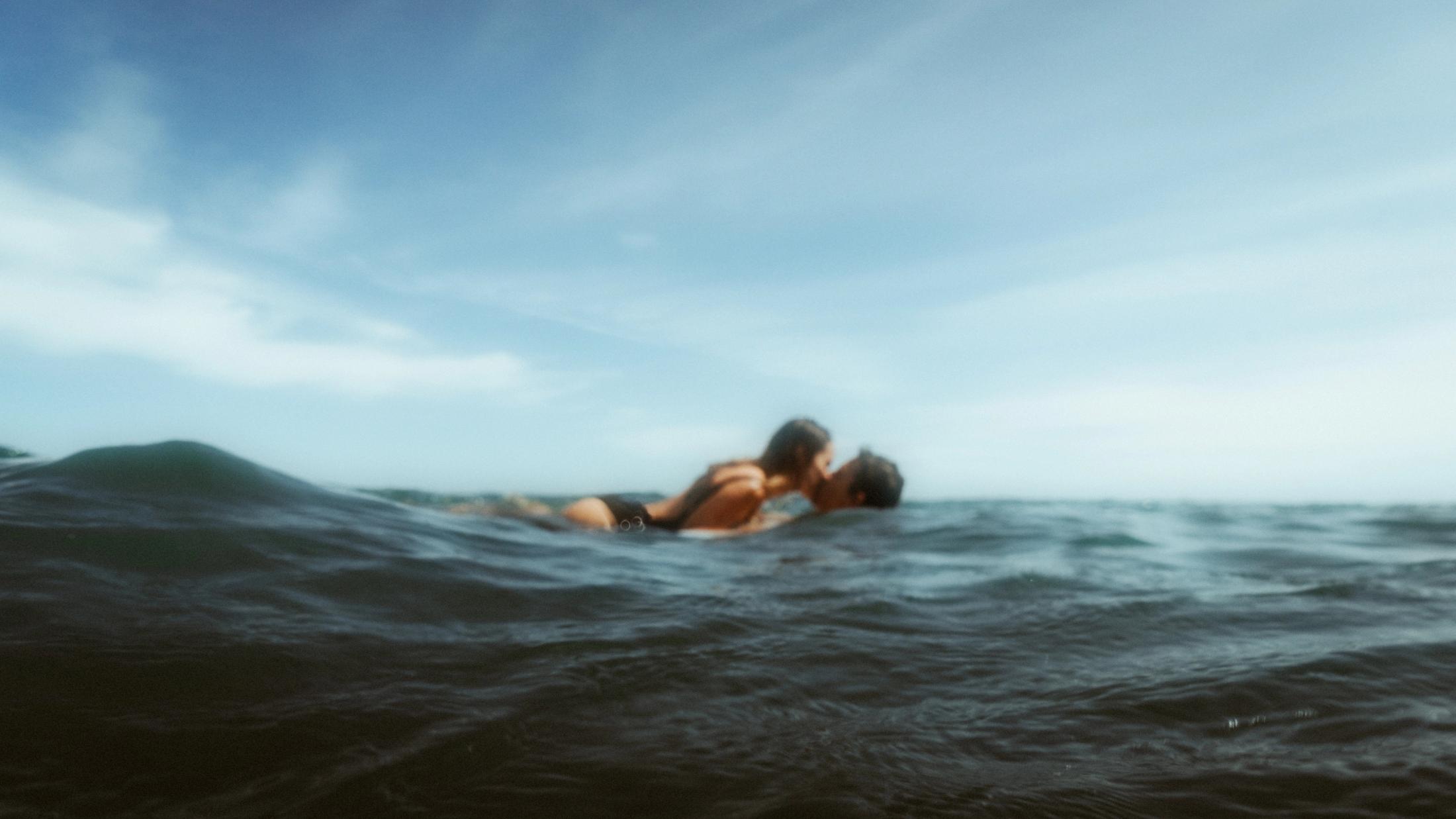 Angela und Ivan küssen sich im offenen Wasser, hinter einer kleinen Welle fotografiert.