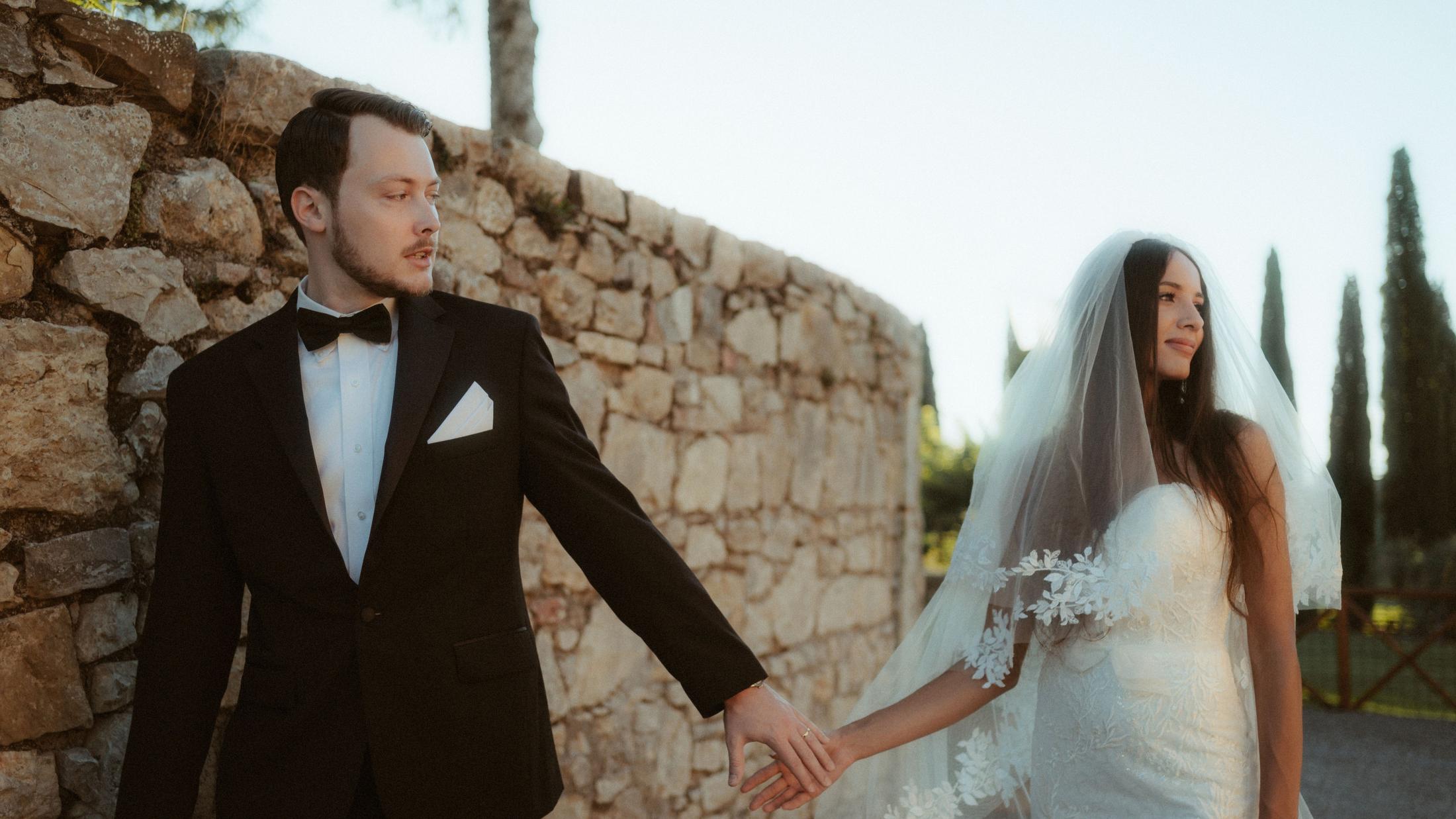 The couple walks hand in hand beside a sunlit stone wall.