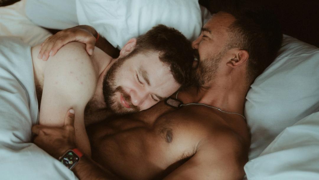 Kris and Ilya lie closely together in an unmade hotel bed.