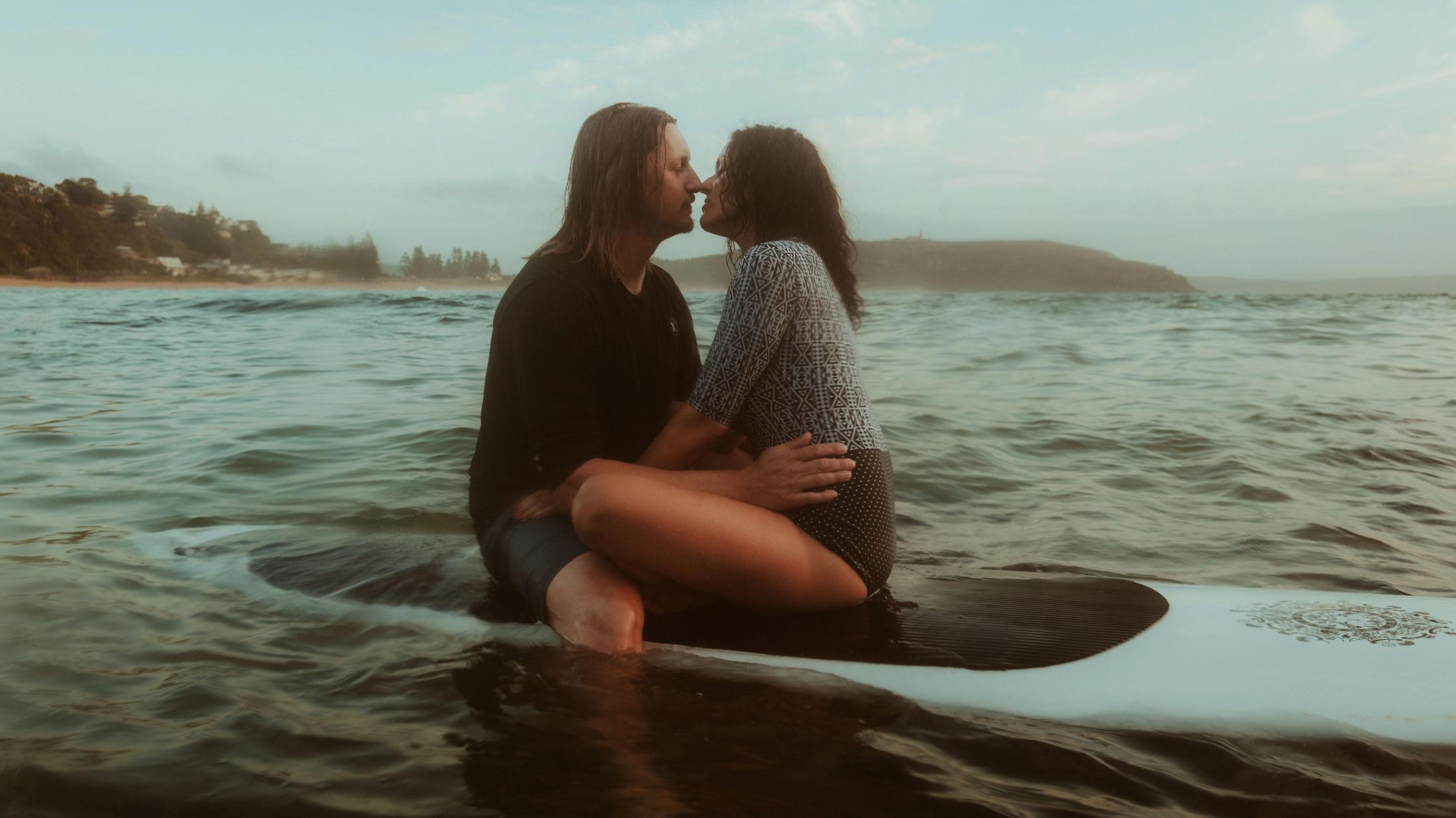 Petra and Roman sit close together on a surfboard in the ocean in soft morning light.