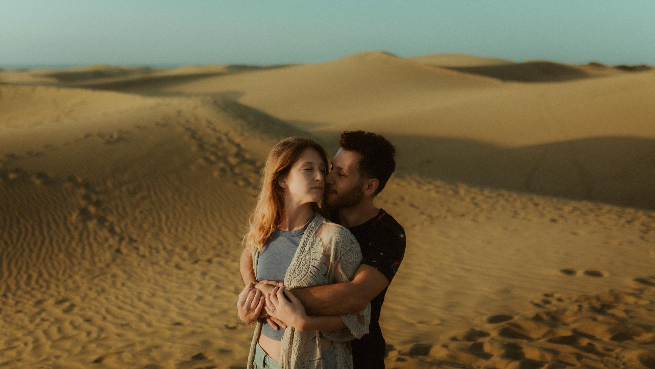 David holding Katja from behind in a wide view of the dunes.