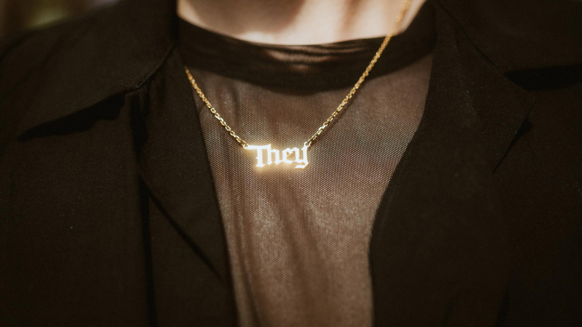 Close view of Noah's gold necklace spelling They over a black shirt.