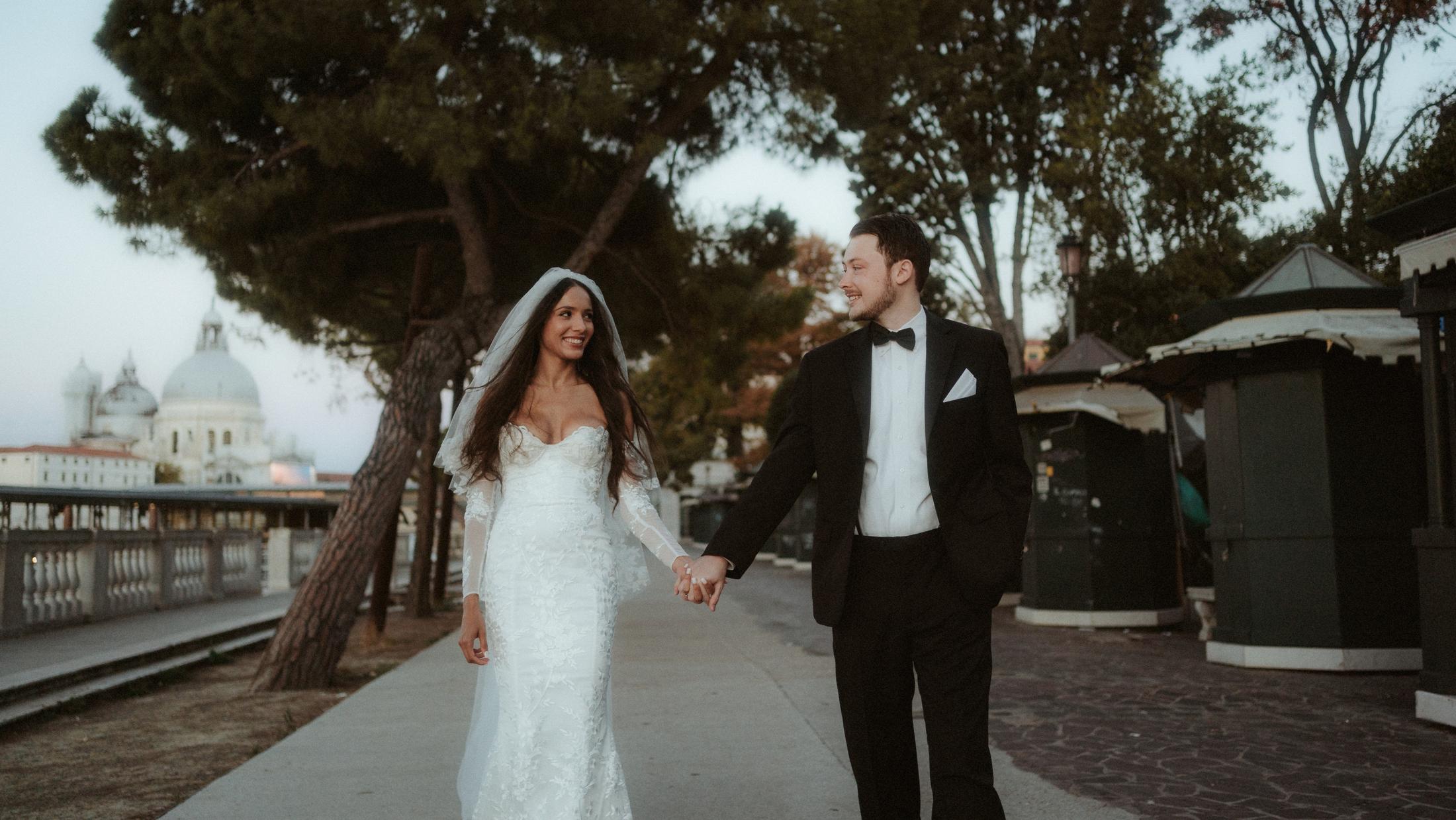 The couple walks along the Venetian waterfront with the lagoon behind them.