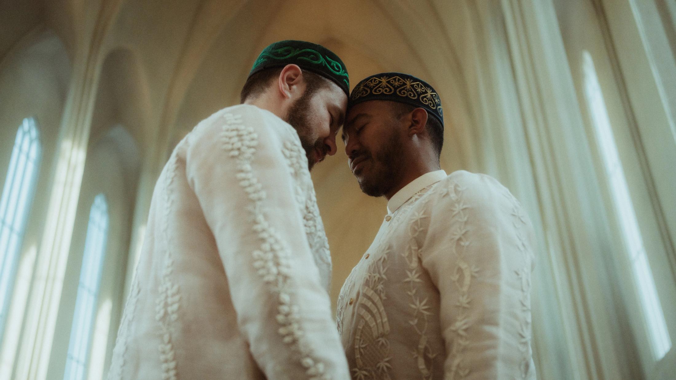 Kris und Ilya lehnen sich drinnen eng aneinander, in einem Barong Tagalog und bestickter kasachischer Kopfbedeckung.