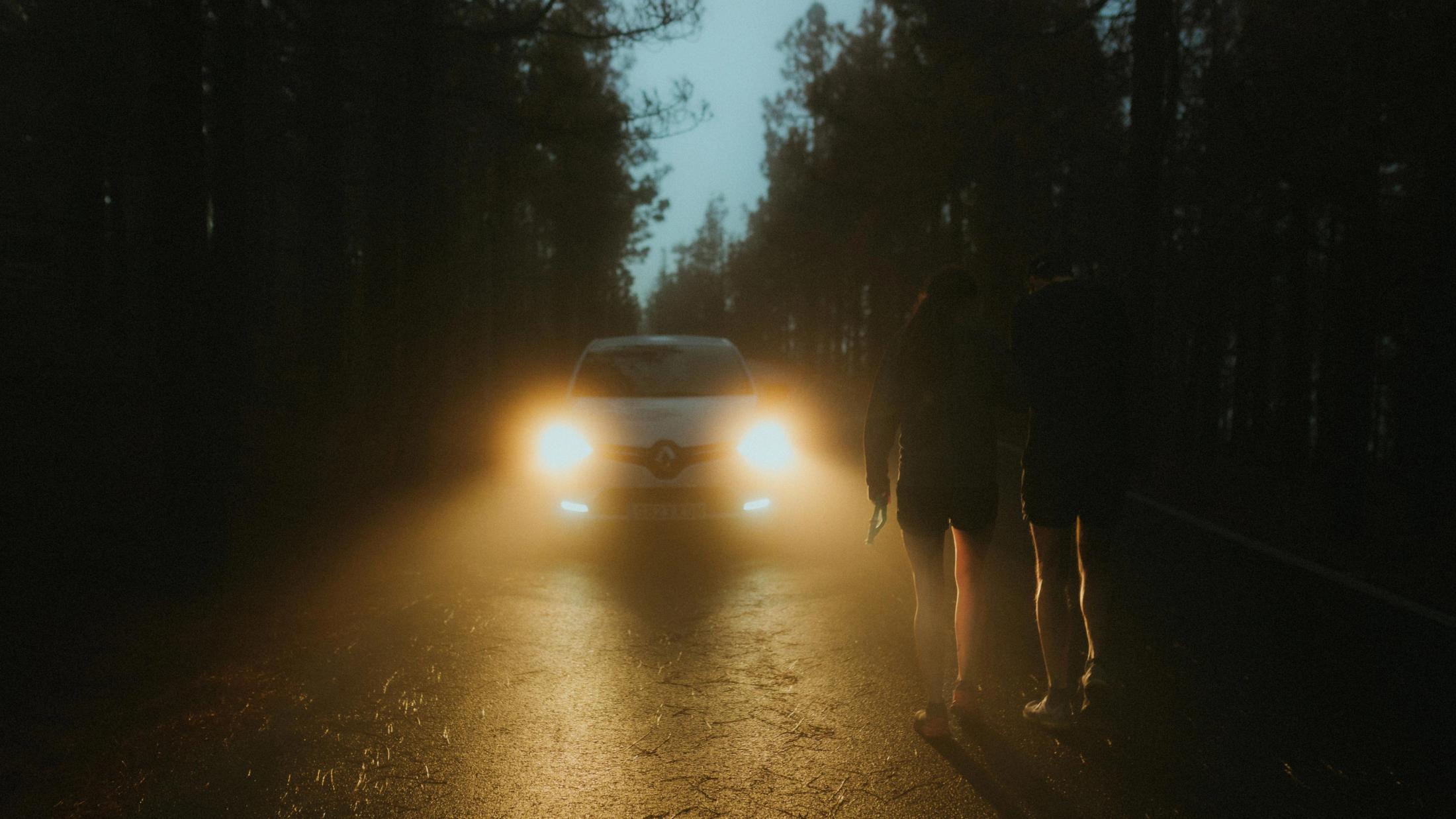 Lisa and Chris stand beside a road at night in the headlights of an approaching car.