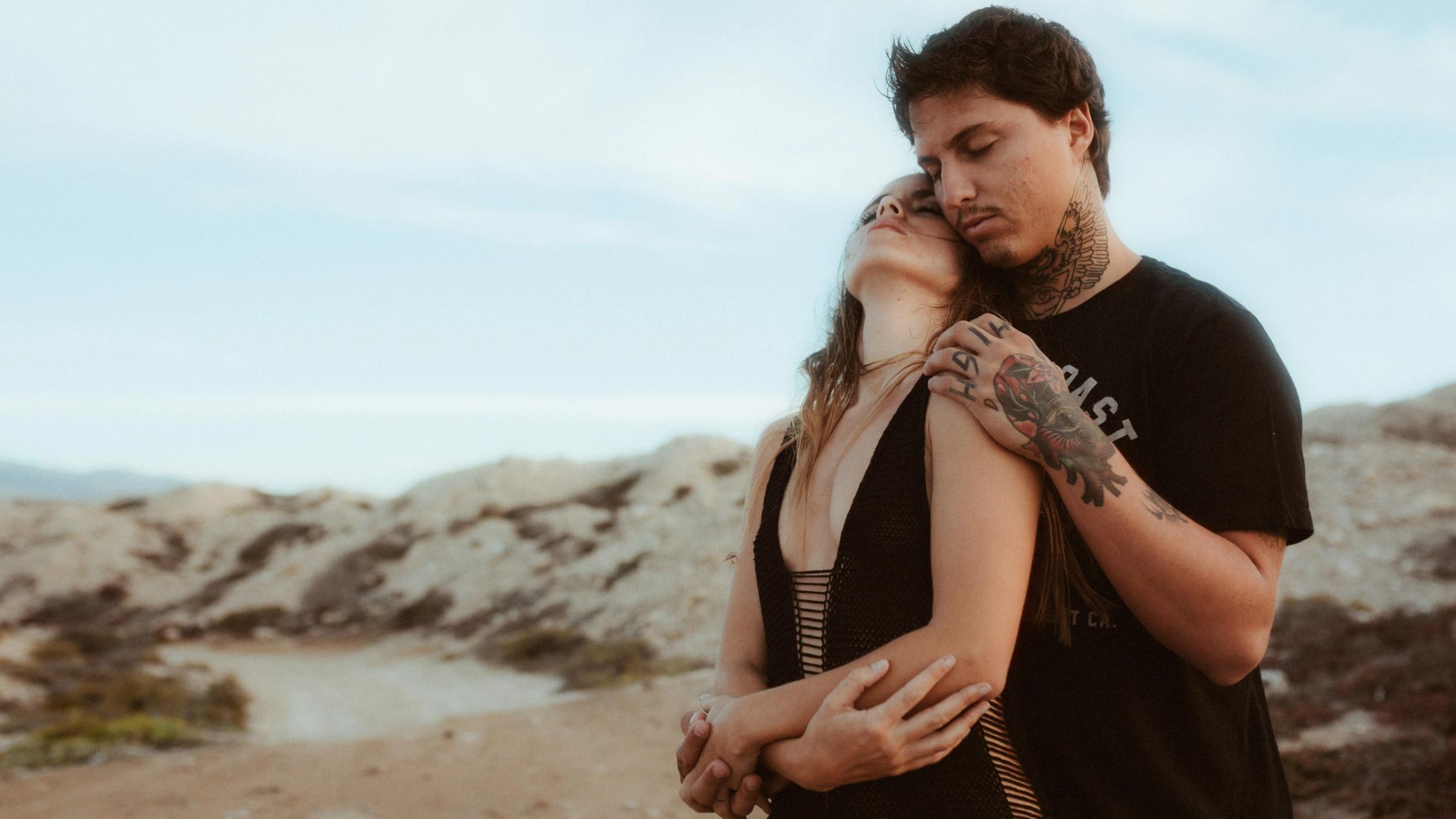 Angela und Ivan umarmen sich auf einer Klippe an der Küste, das Meer im Hintergrund.