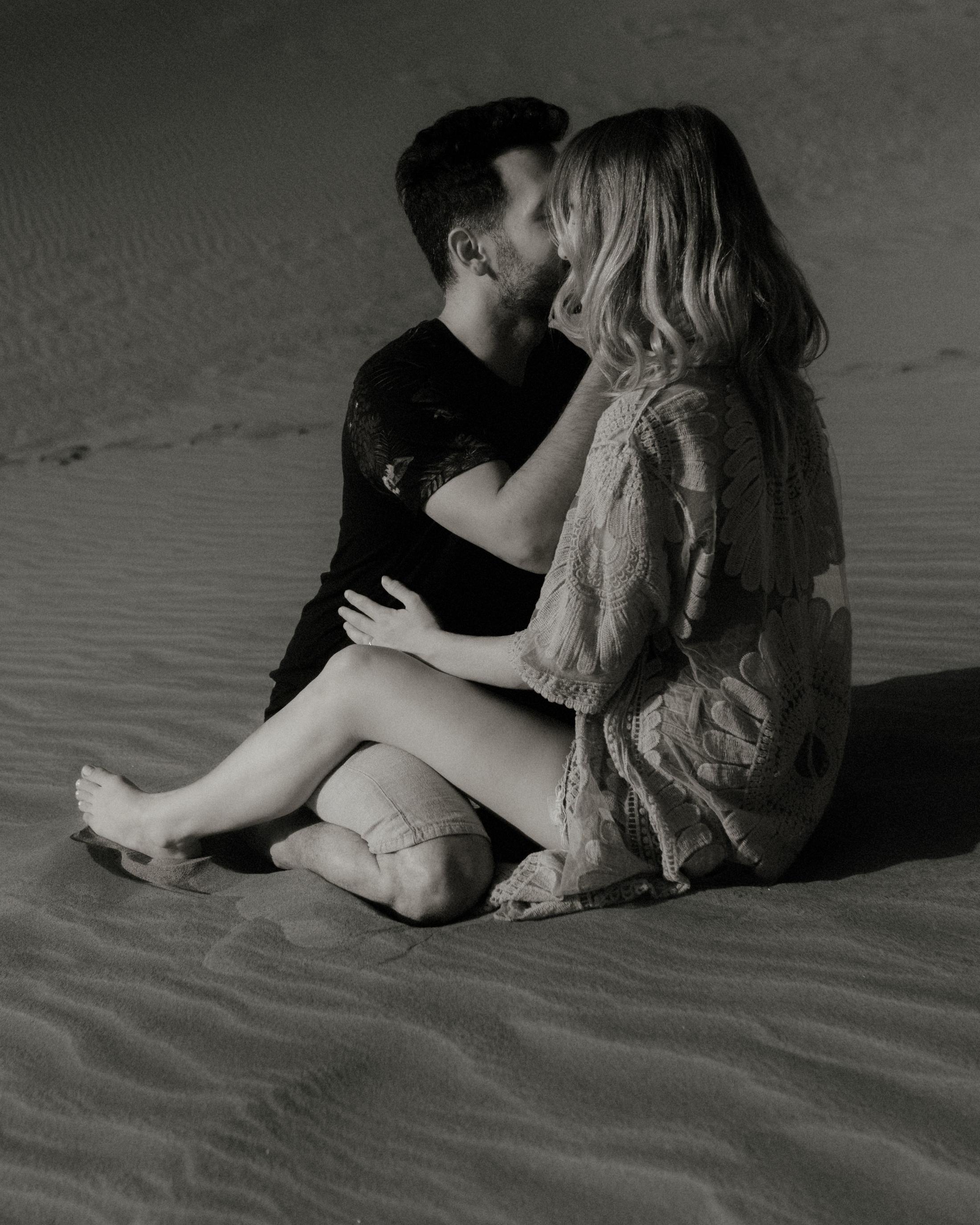 Black-and-white photograph of the couple sitting in the sand and kissing.