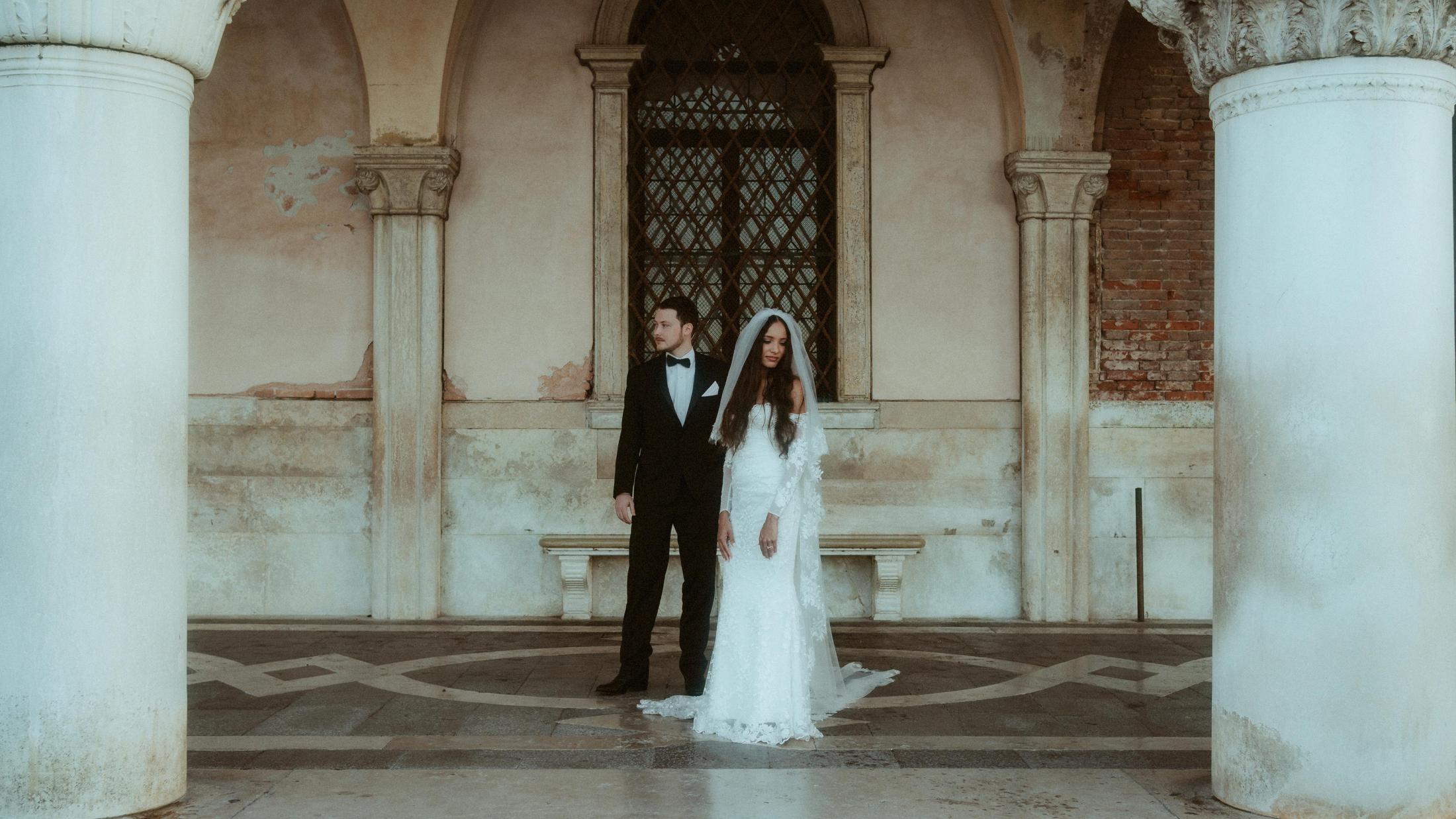 The couple stands between stone columns beneath a Venetian arcade.
