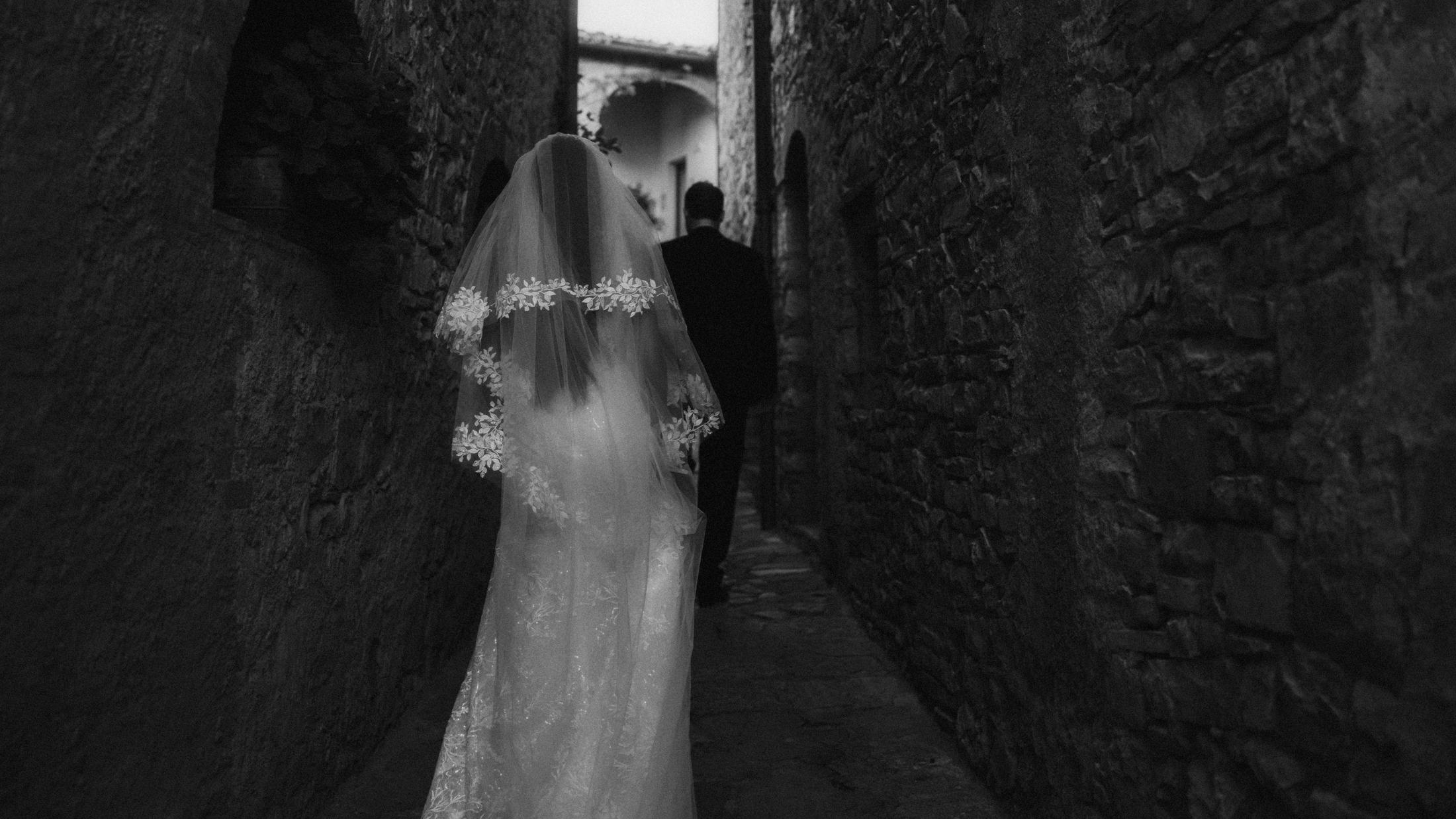 Black-and-white rear view of the couple walking through a narrow stone passage.