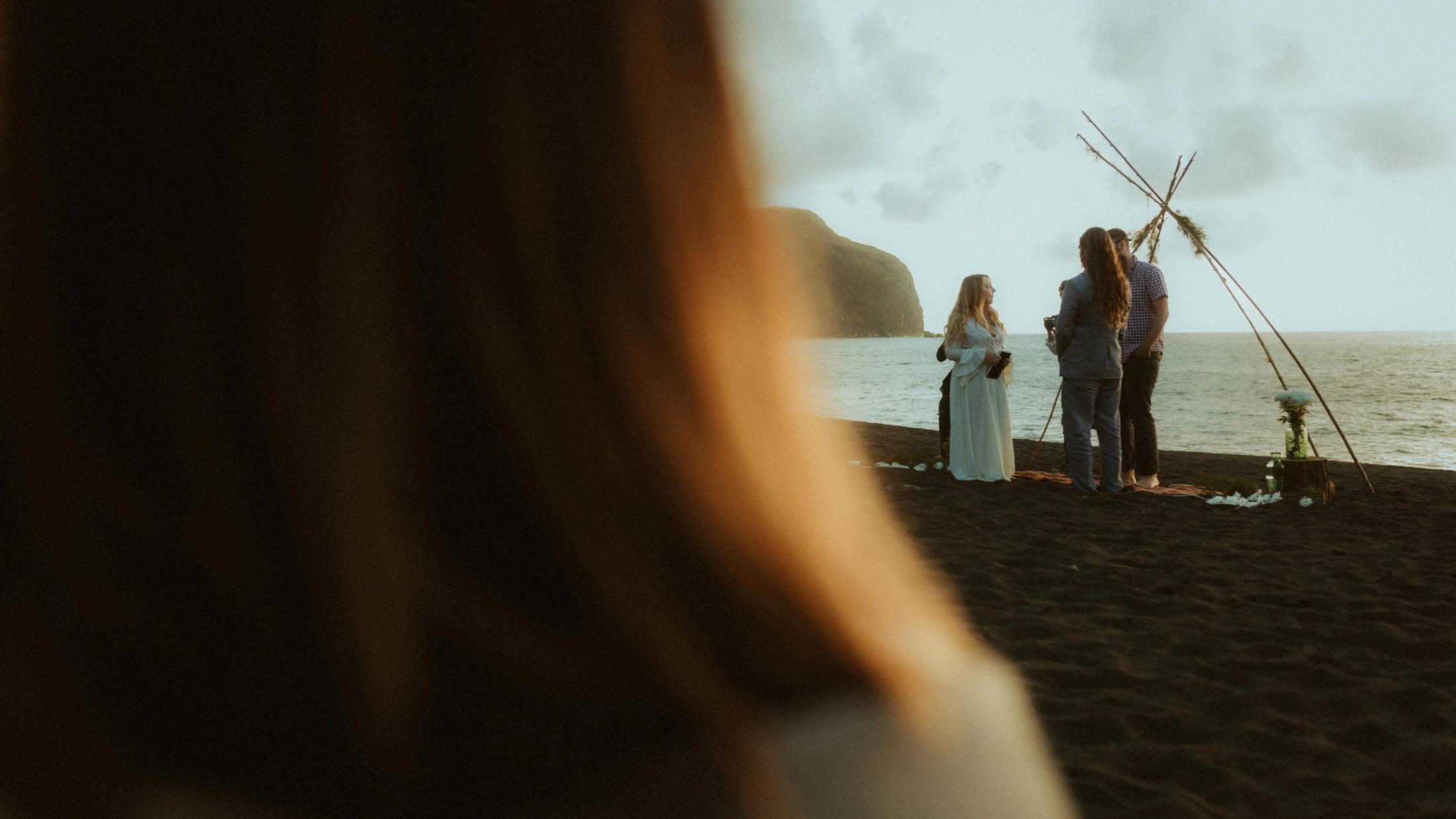 The beach ceremony is photographed past the shoulder of a witness in the foreground.