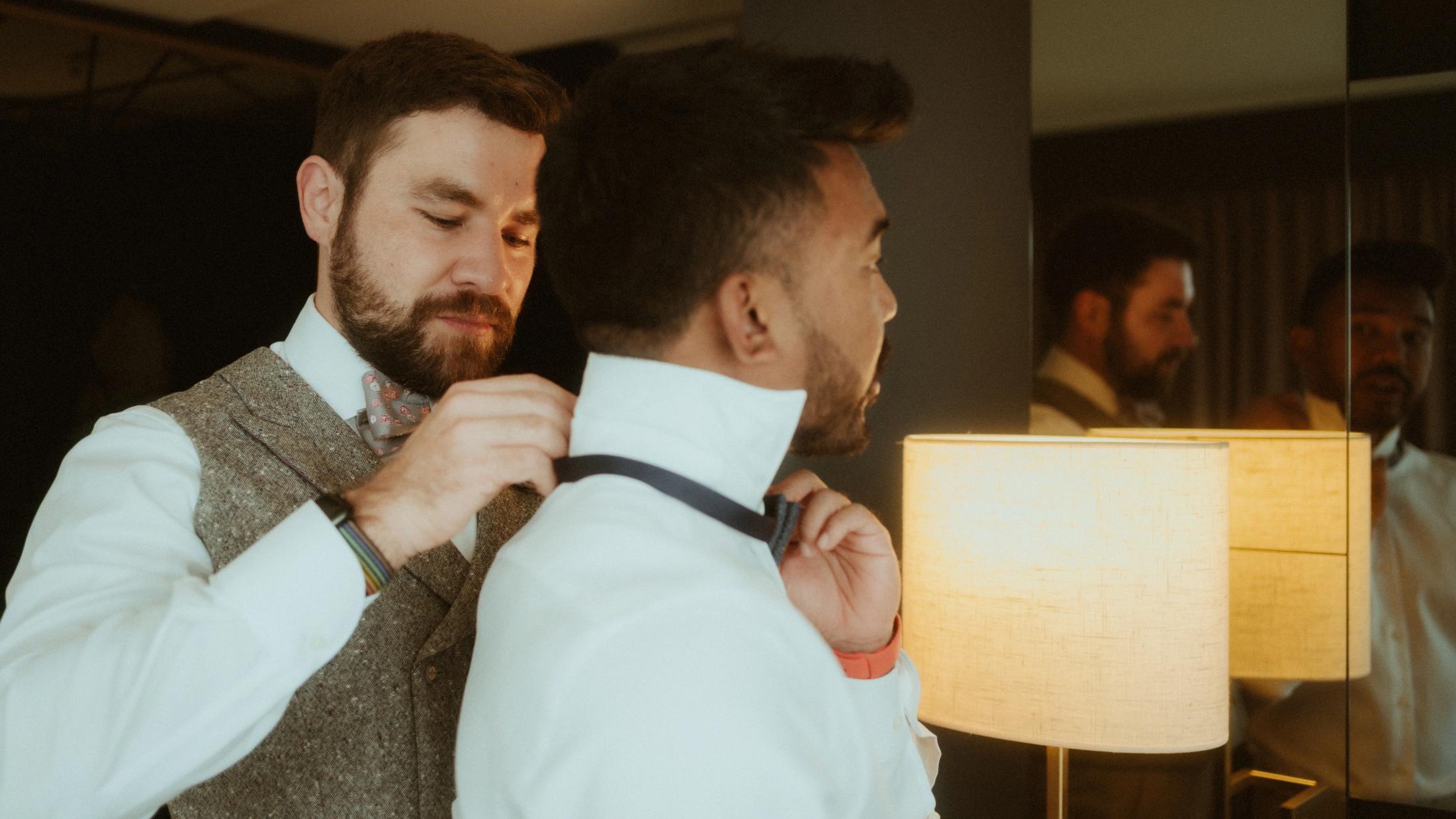 One partner fastens the other's black bow tie beside a hotel lamp.