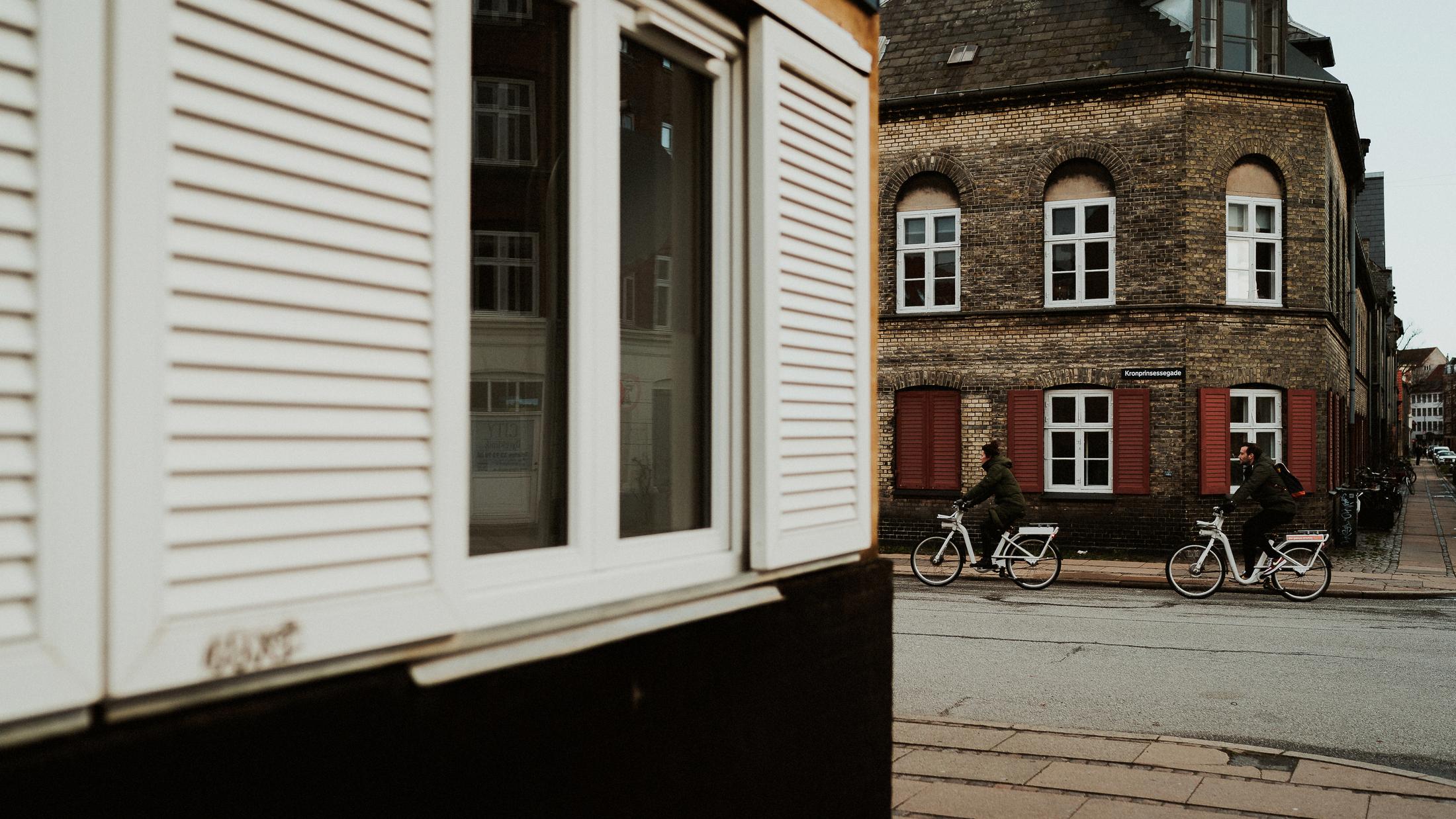 Two cyclists pass a brick building, framed by a white bay window.