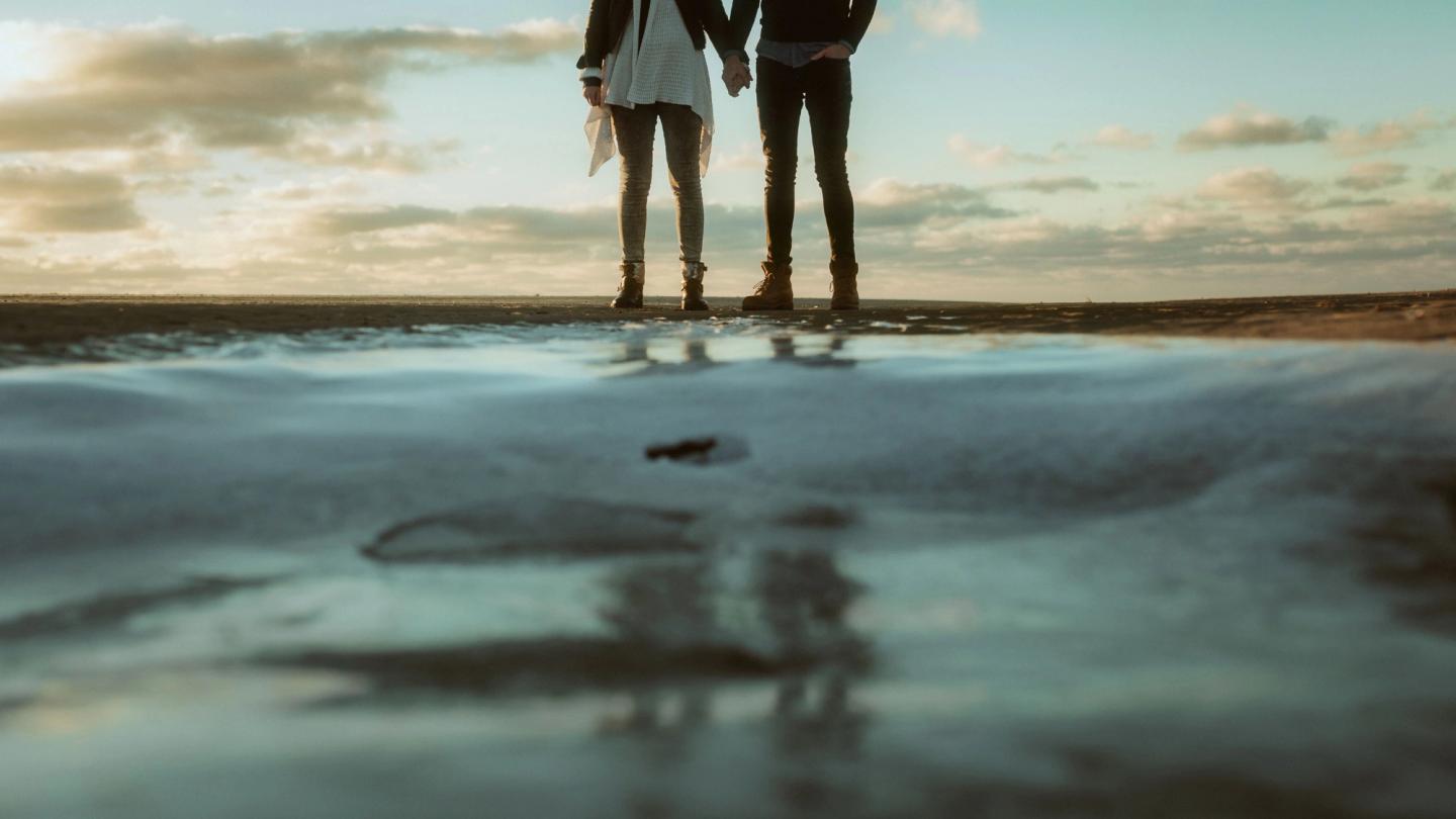 Hanna and Henrik stand hand in hand on the beach, reflected in a shallow pool of water.
