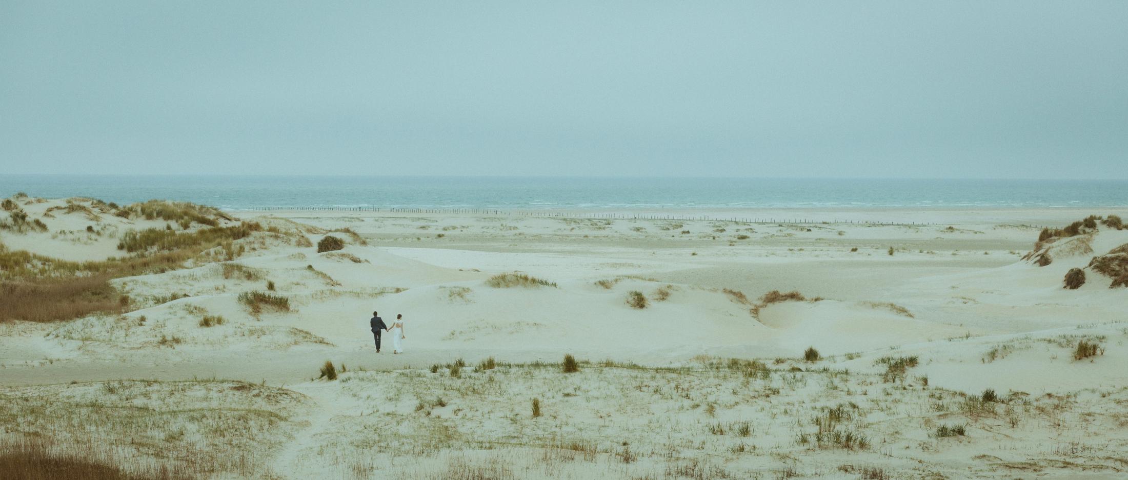Nina und Torben erscheinen als kleine Figuren zwischen hellen Dünen mit Blick auf das ferne Meer.