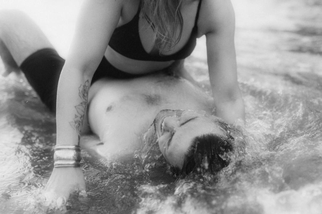 Schwarz-weiß Detailaufnahme von Badeanzug und Tattoo einer Frau, der Kopf ihres Partners ruht darunter im Wasser, Hafenliebe