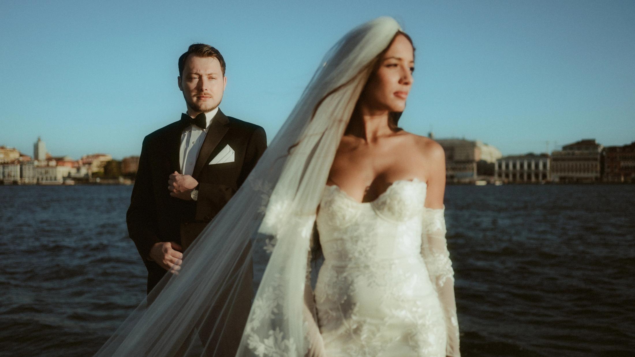 Cinematic waterfront portrait with the bride in a flowing veil and her partner standing behind her.