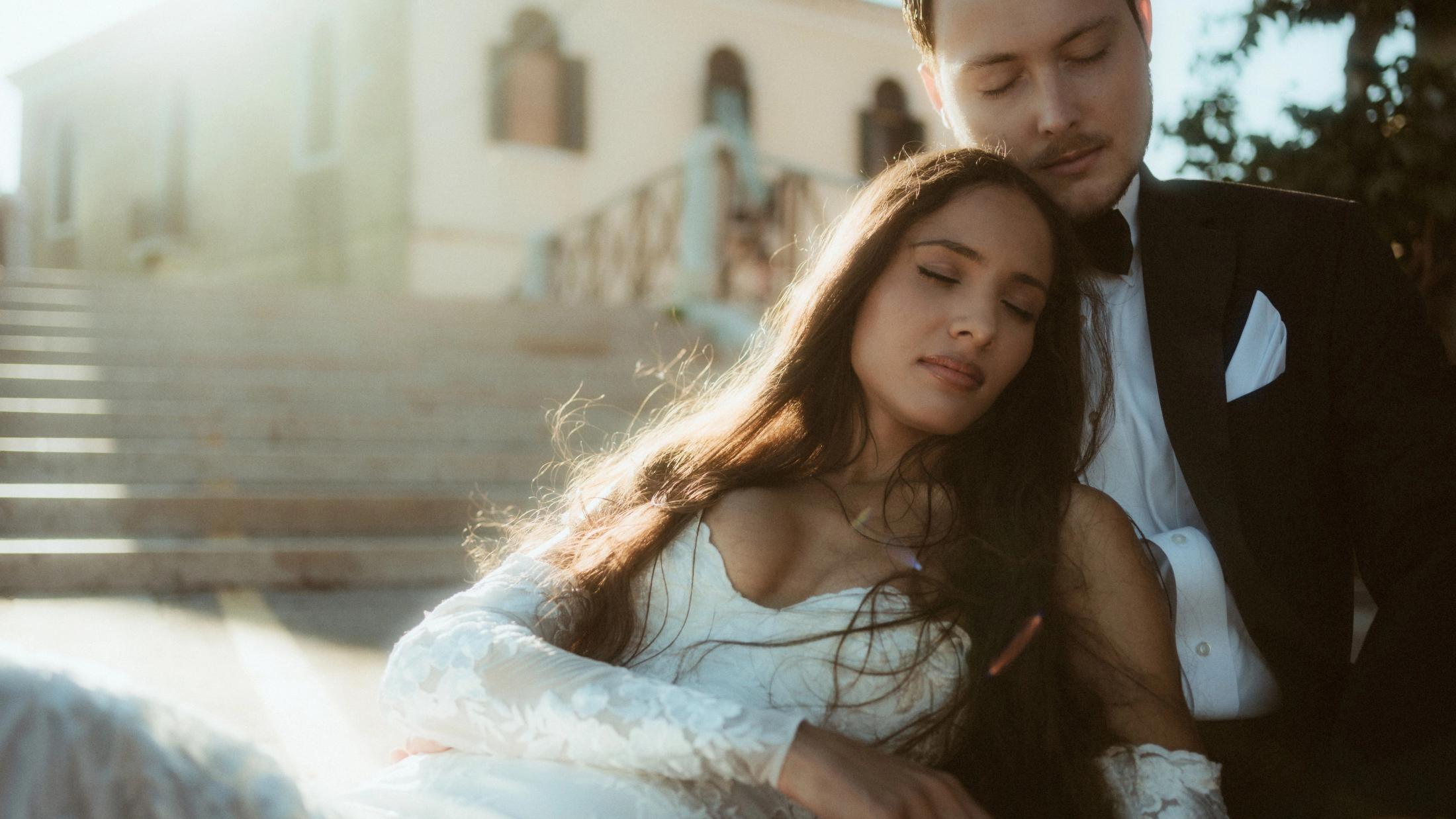 The bride rests against her partner while they sit together on sunlit steps.