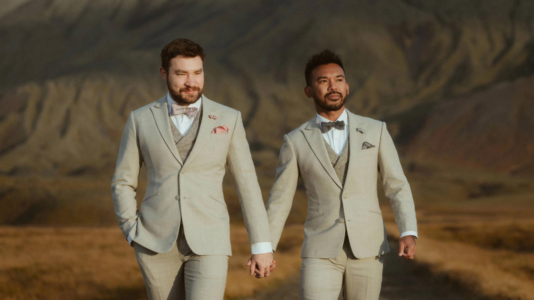 Kris und Ilya gehen in Anzügen auf die Kamera zu, hinter ihnen ein dunkler Berg.