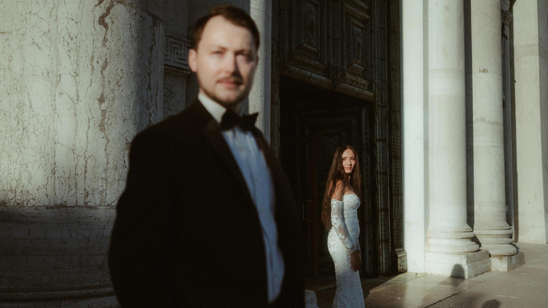 The groom is out of focus in front while the bride stands sharply in a church doorway.