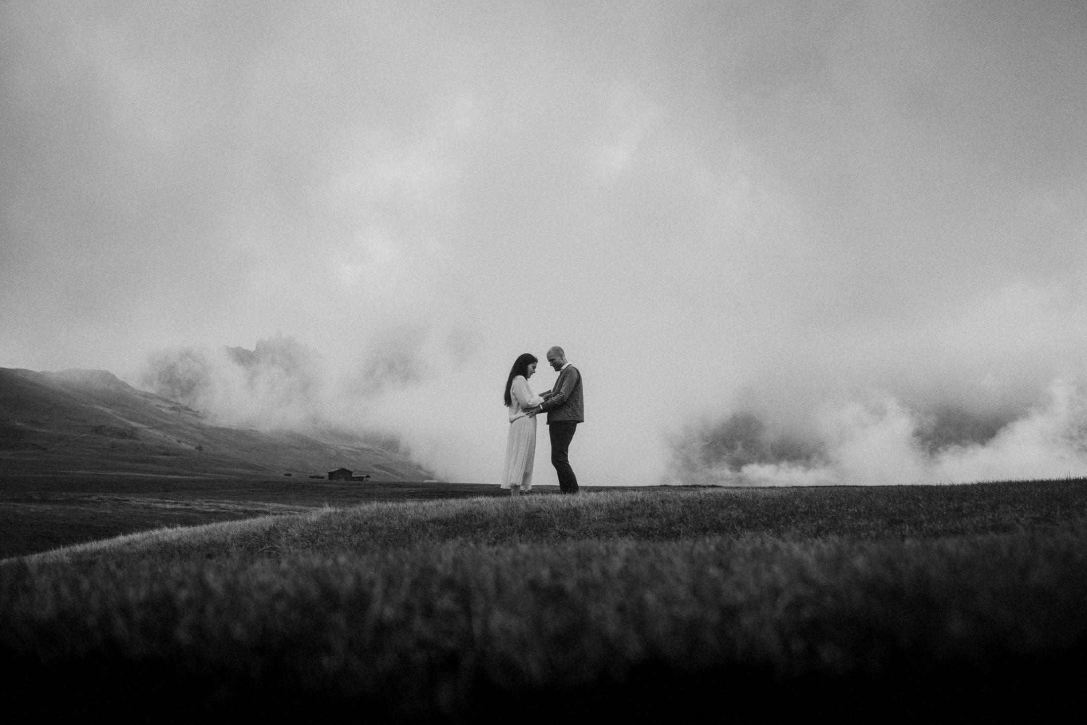 The couple embracing with mountains and moving clouds behind them.