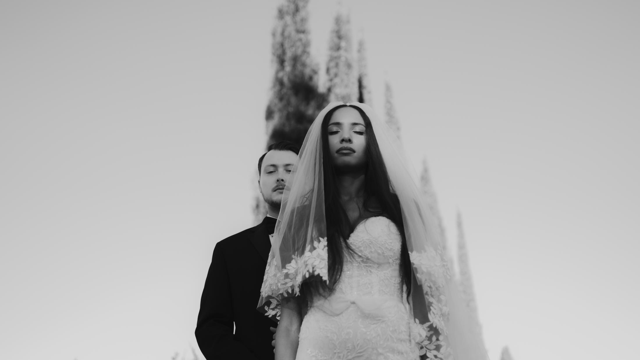Low-angle black-and-white portrait of the couple beneath blurred cypress trees.