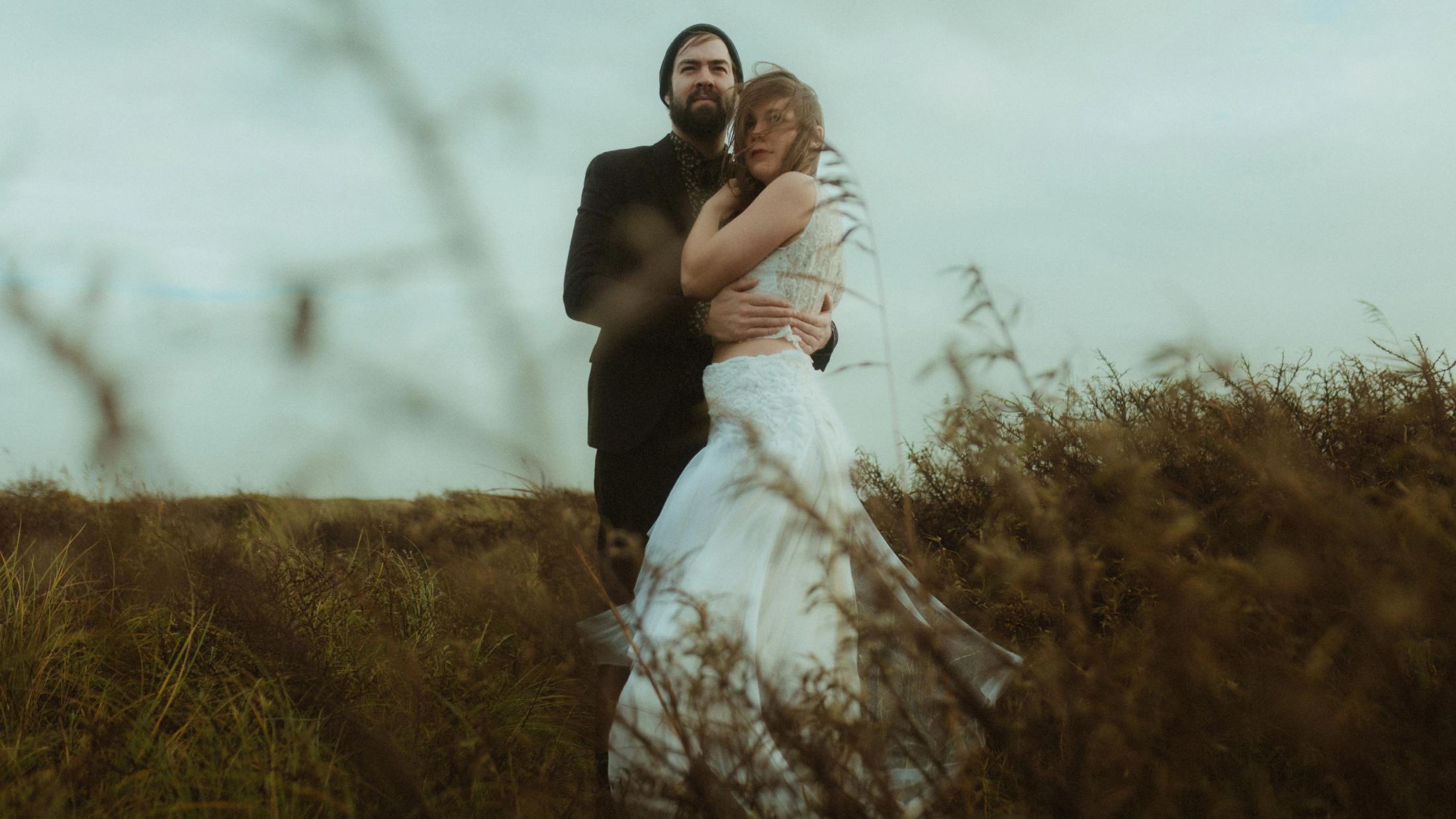 Anni and Daniel hold each other behind blurred dune grass.
