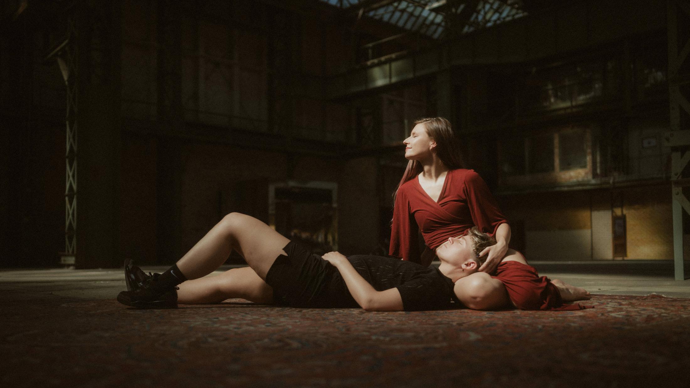 Noah resting with their head in Michèle's lap inside the dim industrial hall.