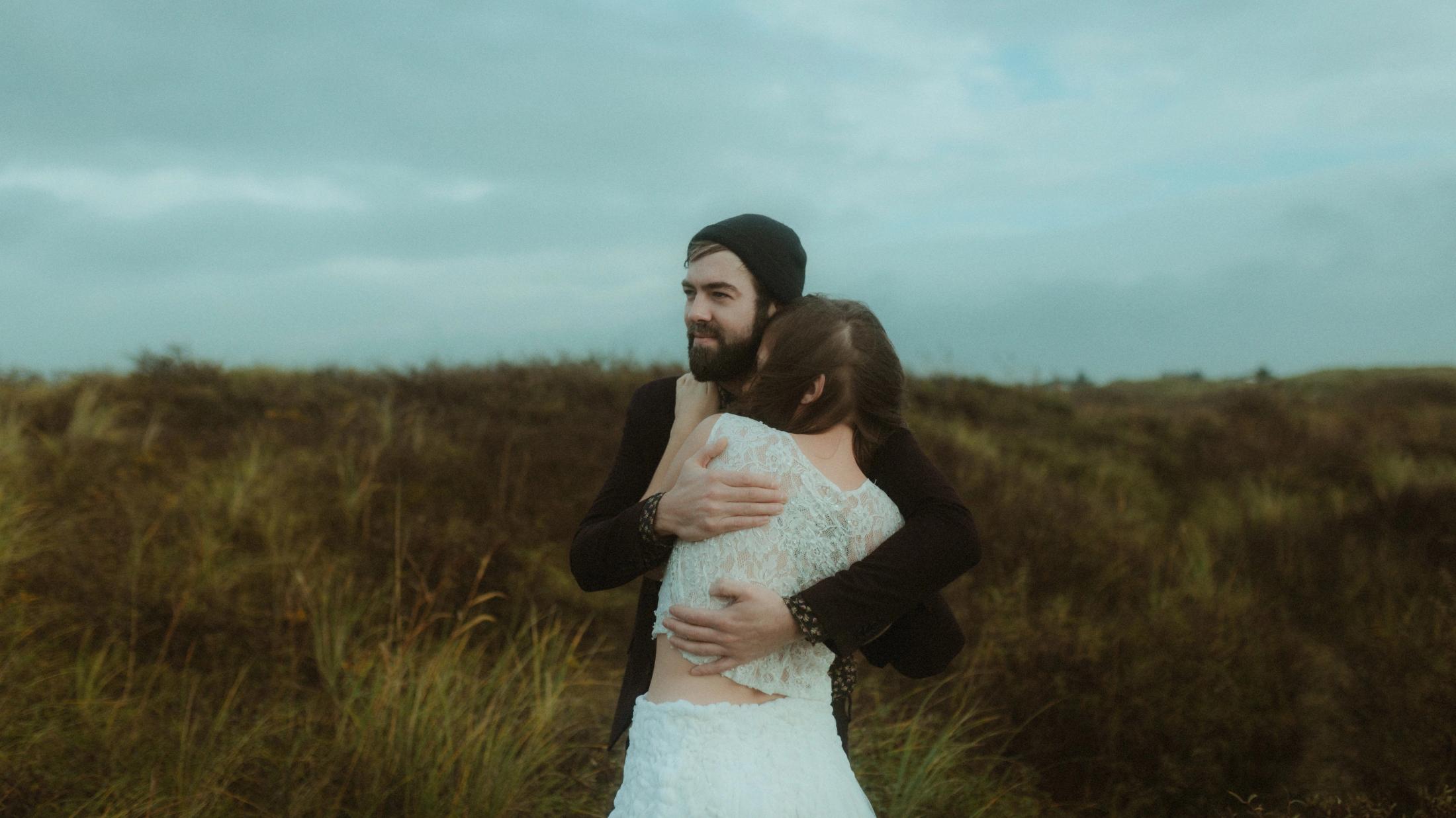 The couple hold each other in the open grassland under a grey sky.