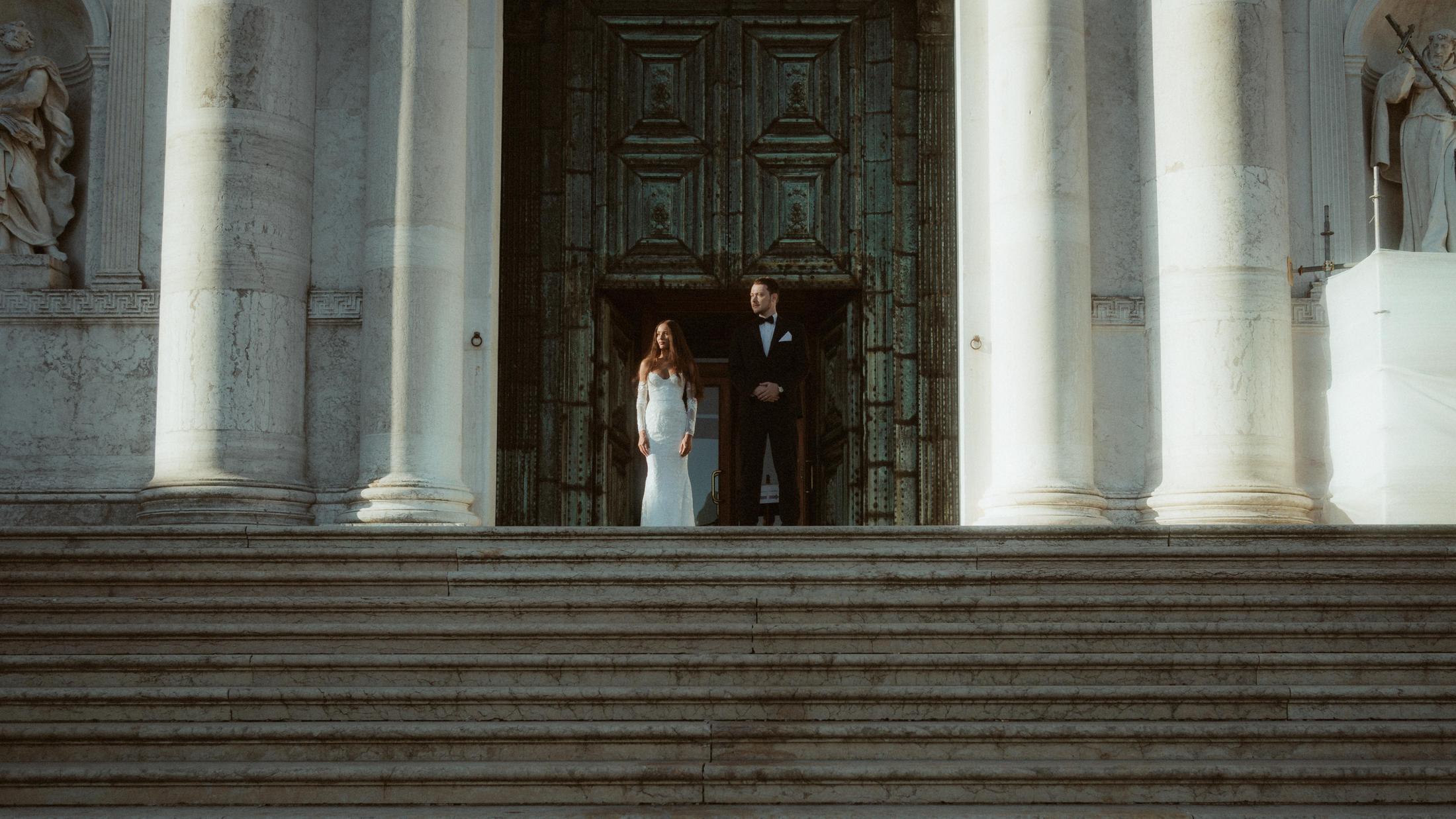 The couple stands in deep shadow inside a vast church doorway above broad stone steps.