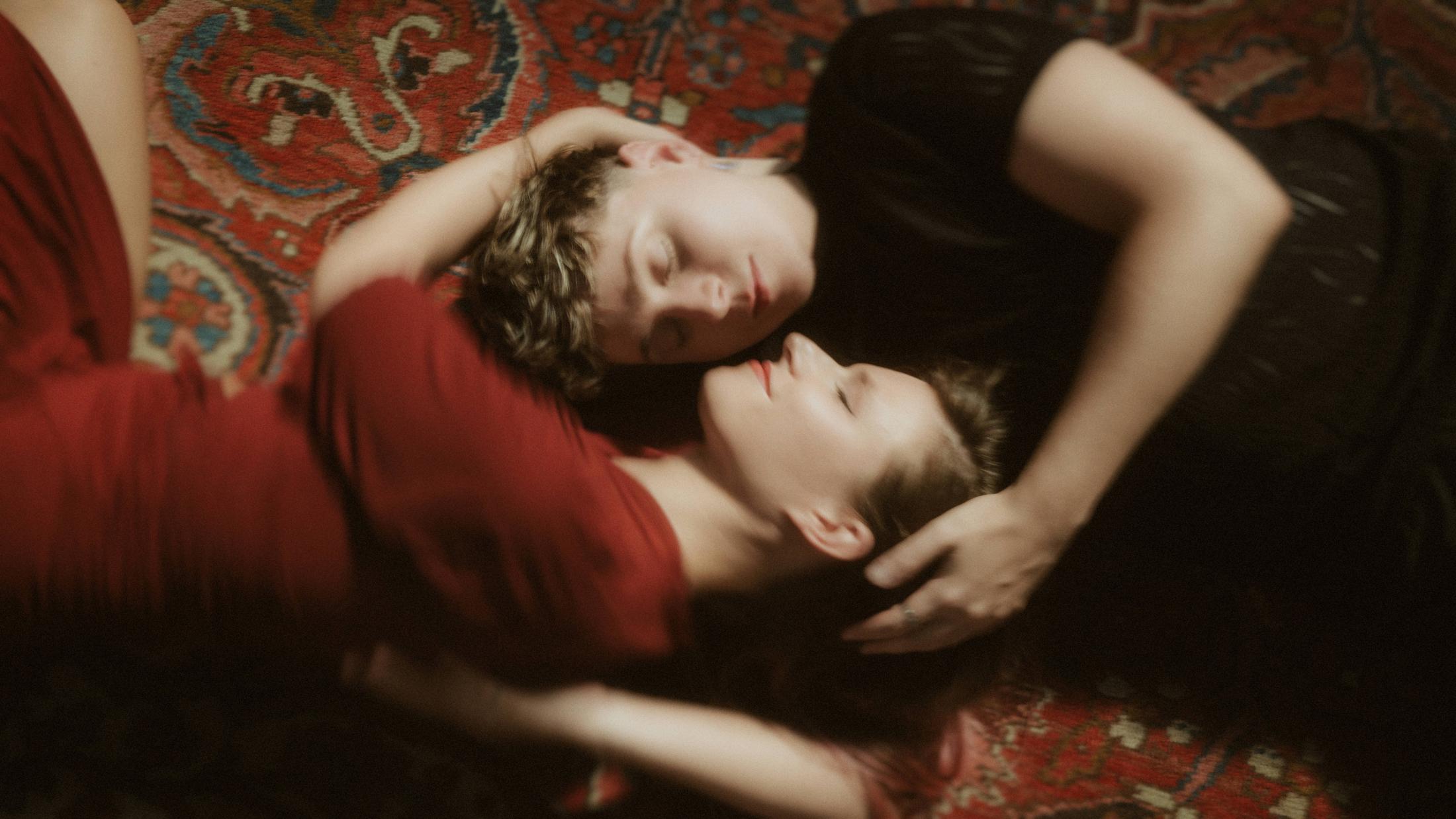 Noah and Michèle lying face to face on the red carpet inside the industrial hall.