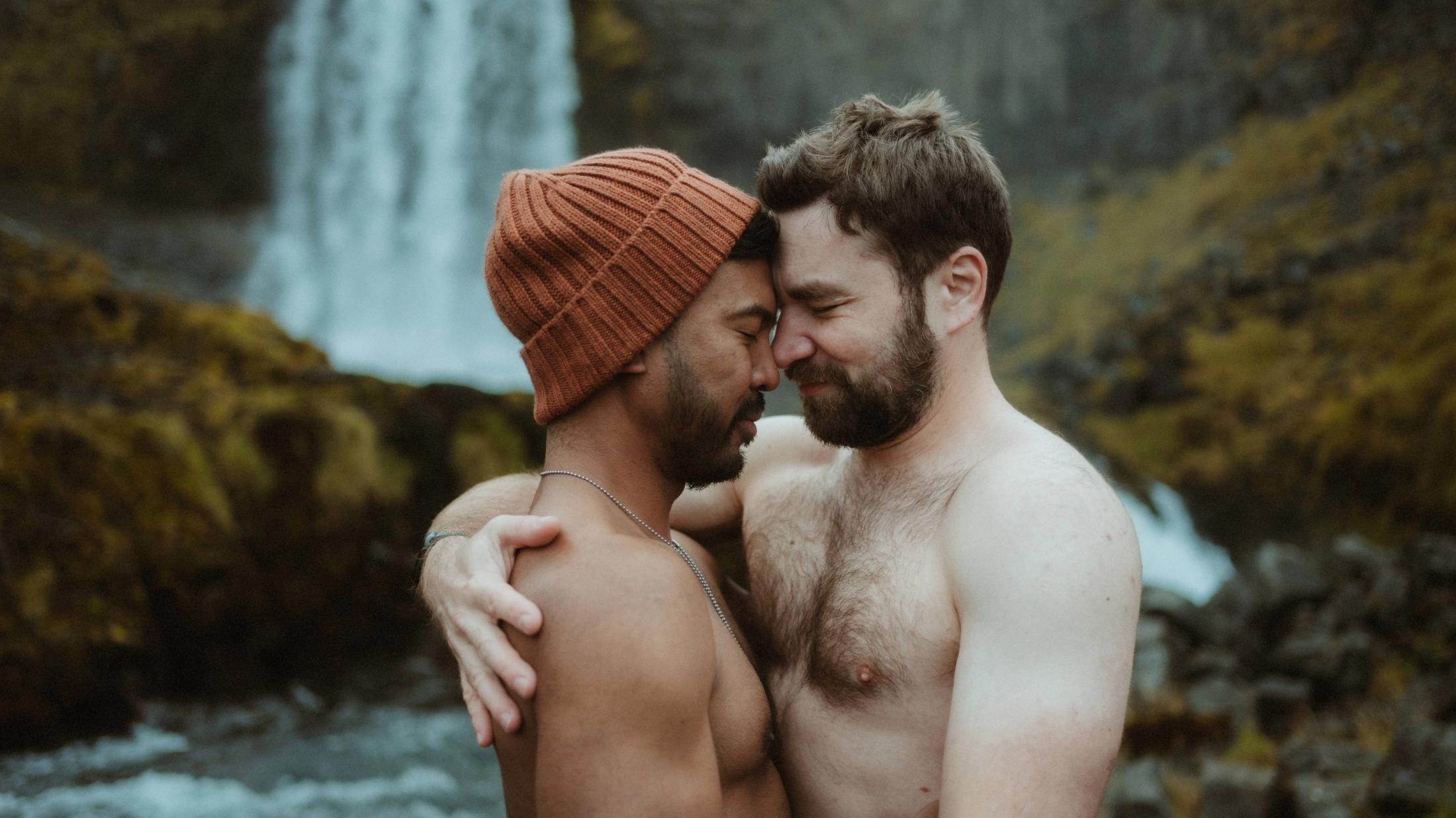 Kris und Ilya umarmen sich mit nacktem Oberkörper vor dem Wasserfall.