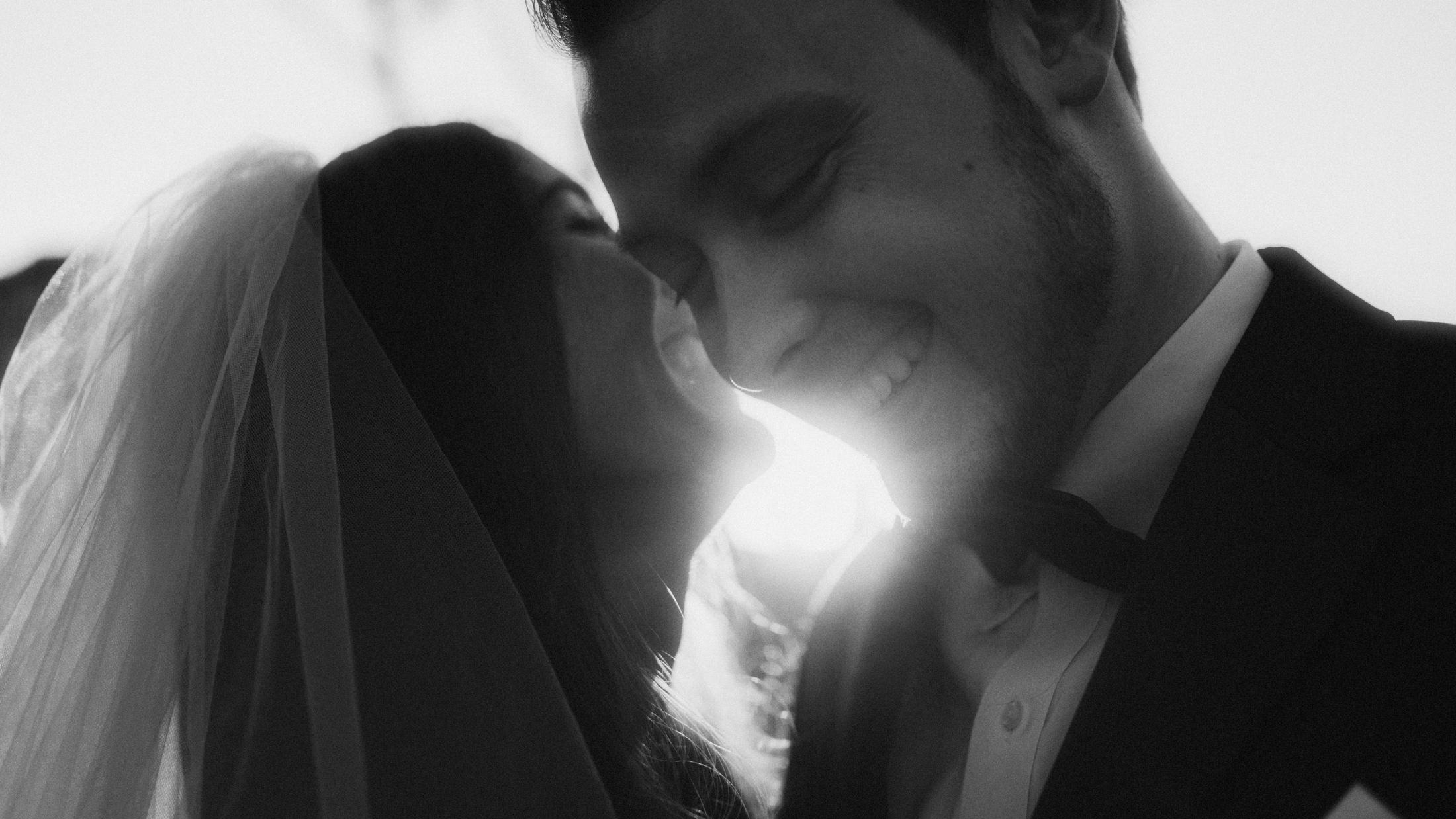 Backlit black-and-white close-up of the couple touching faces beneath the veil.