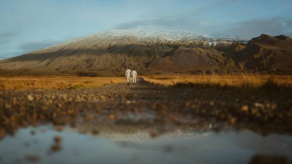 Kris and Ilya walk through an Icelandic valley beneath a snow-covered ridge, reflected in shallow water.