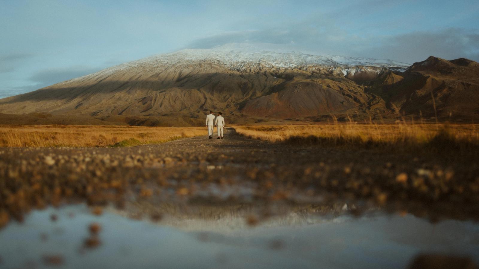 Kris and Ilya walk through an Icelandic valley beneath a snow-covered ridge, reflected in shallow water.