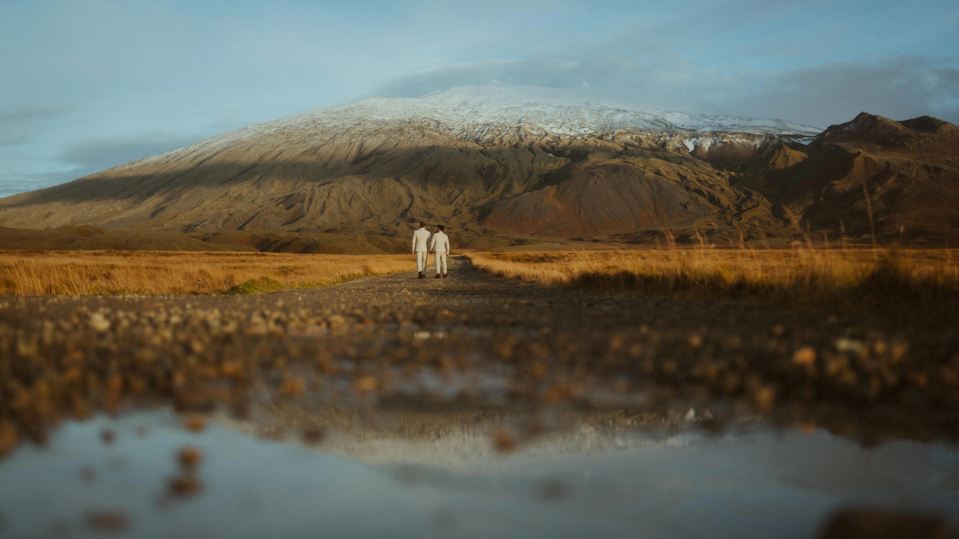 Kris and Ilya walk through an Icelandic valley beneath a snow-covered ridge, reflected in shallow water.