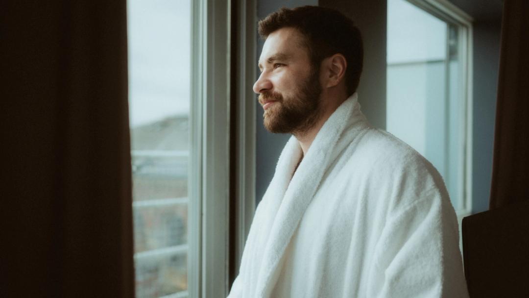 Ilya looks out of the hotel window in a white robe.