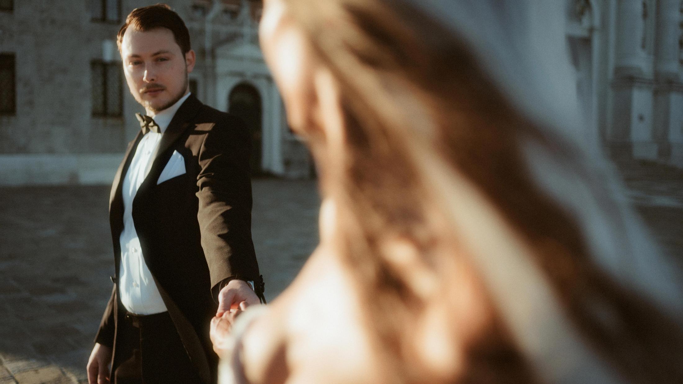The groom offers his hand while the bride crosses the foreground out of focus.