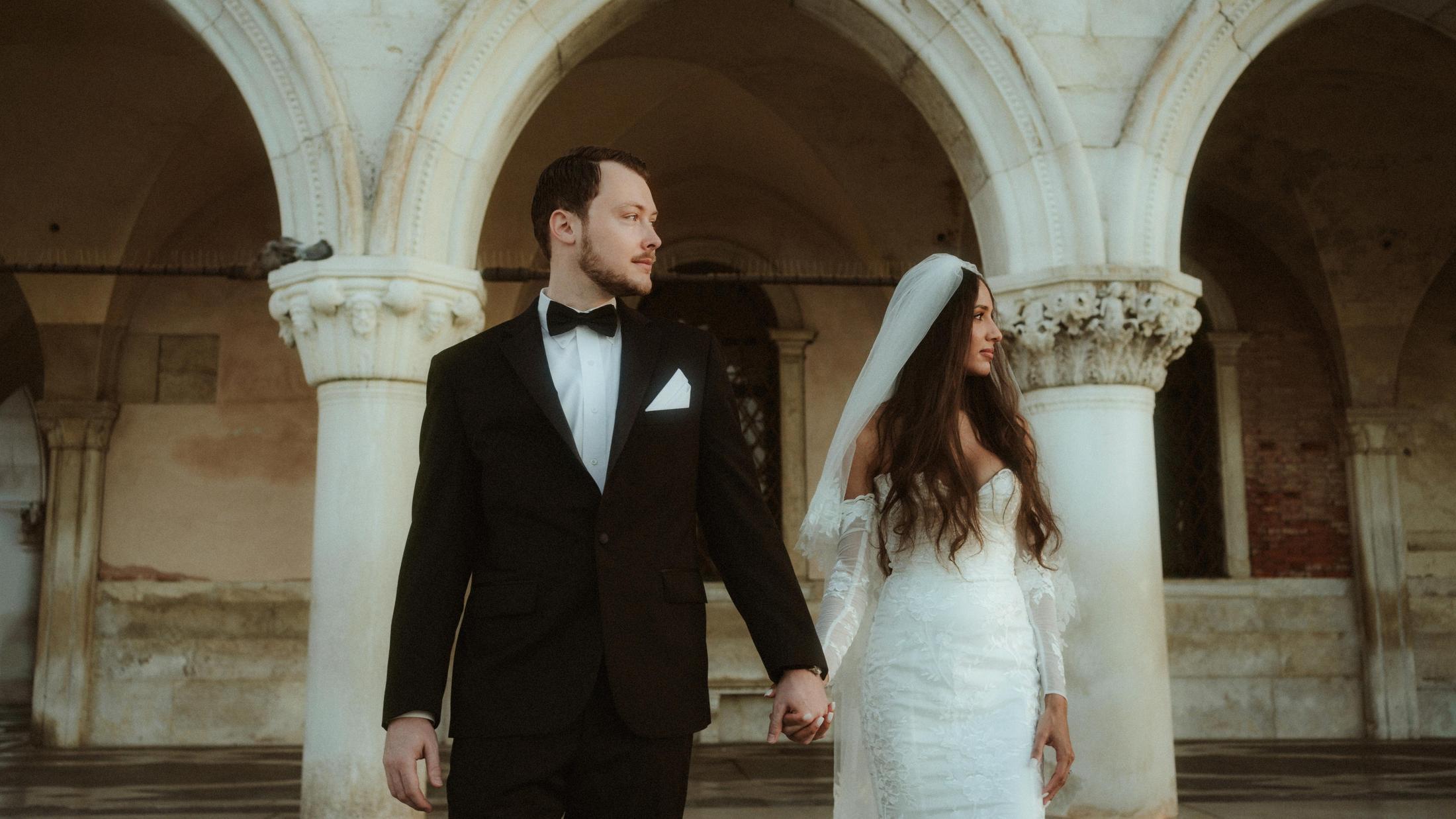 The couple holds hands while looking in opposite directions beneath the arches.