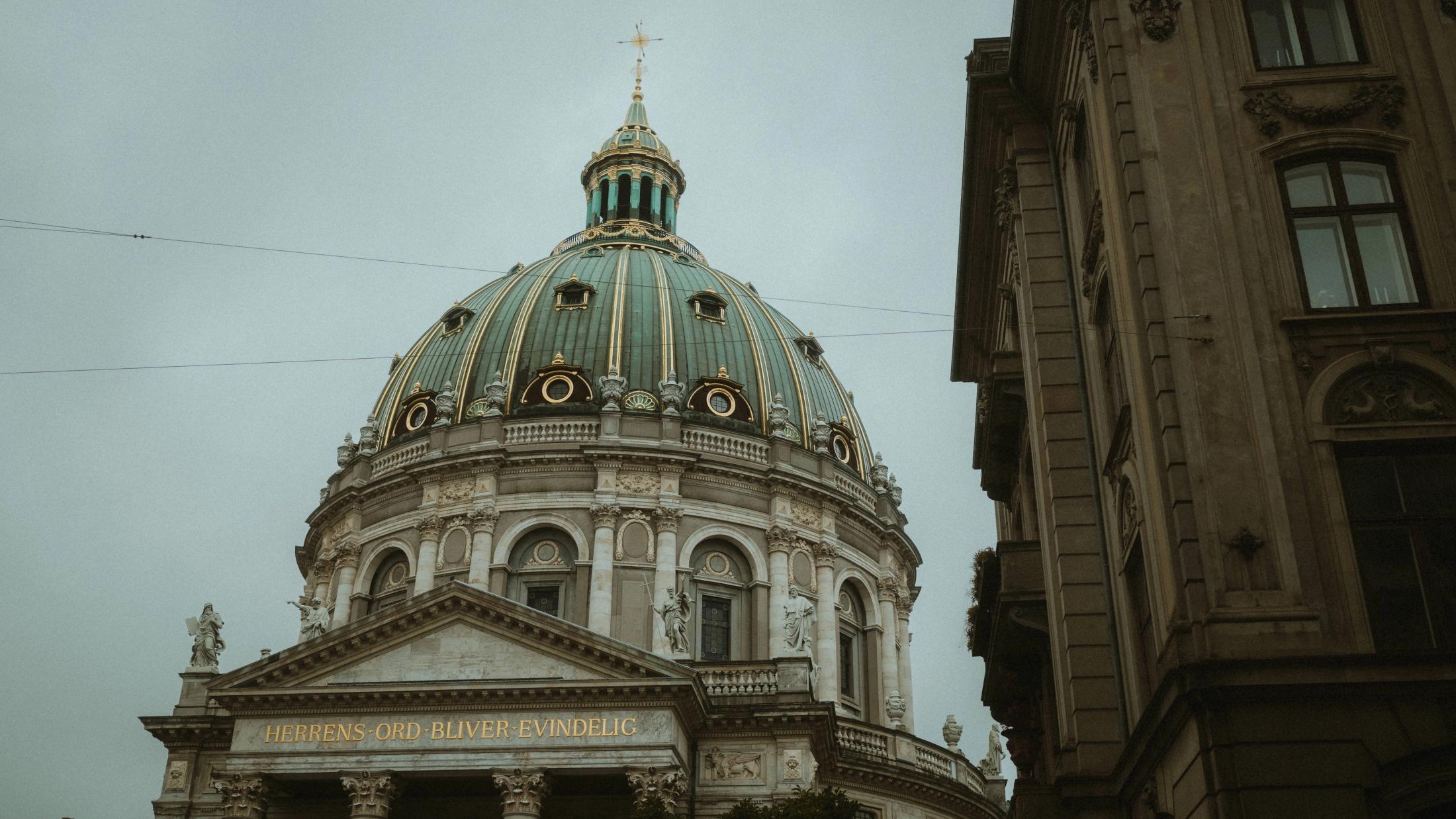 A green copper dome rises beside a dark stone facade beneath an overcast sky.